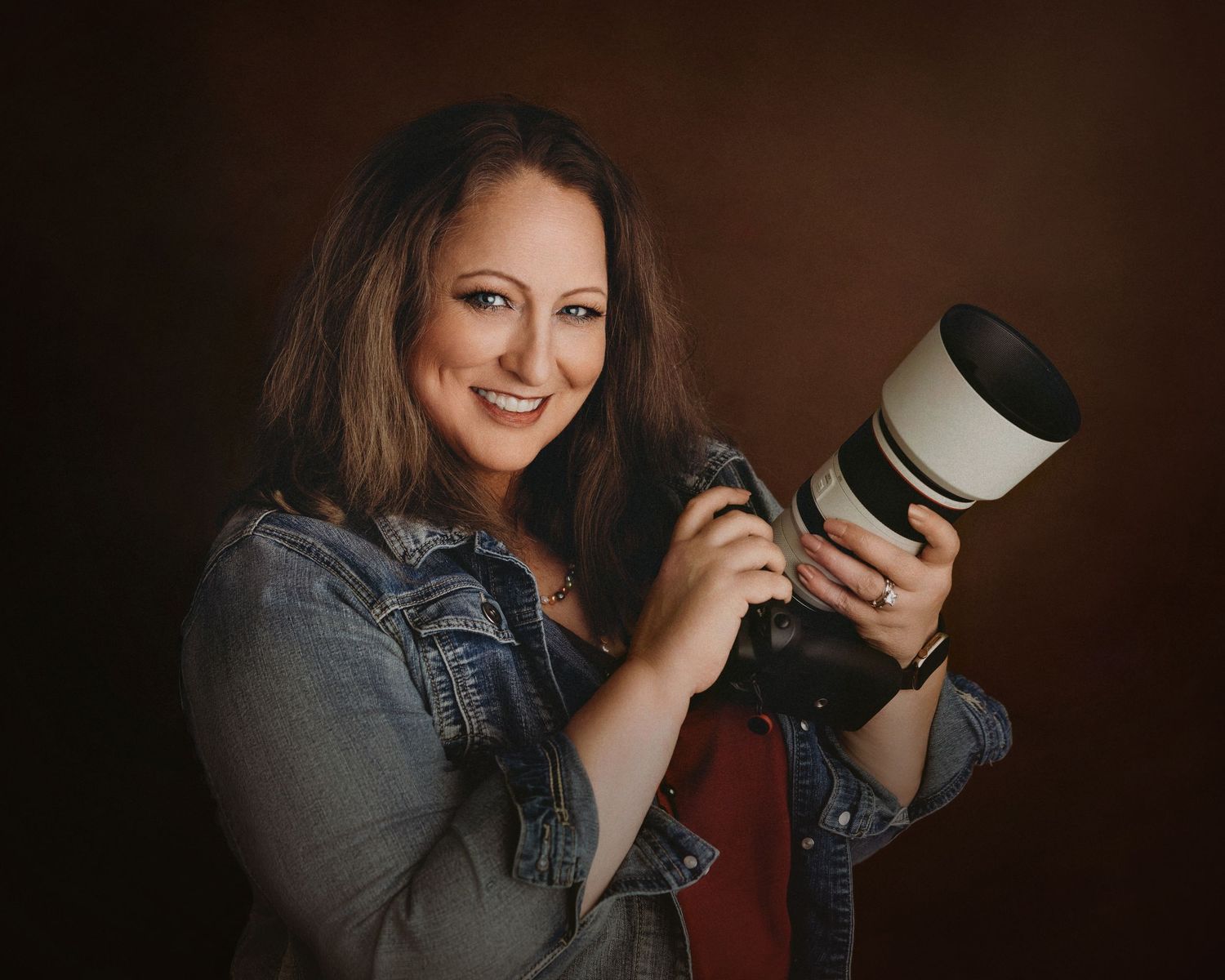 Professional photographer in denim shirt holds camera lens while working in dark studio setting.