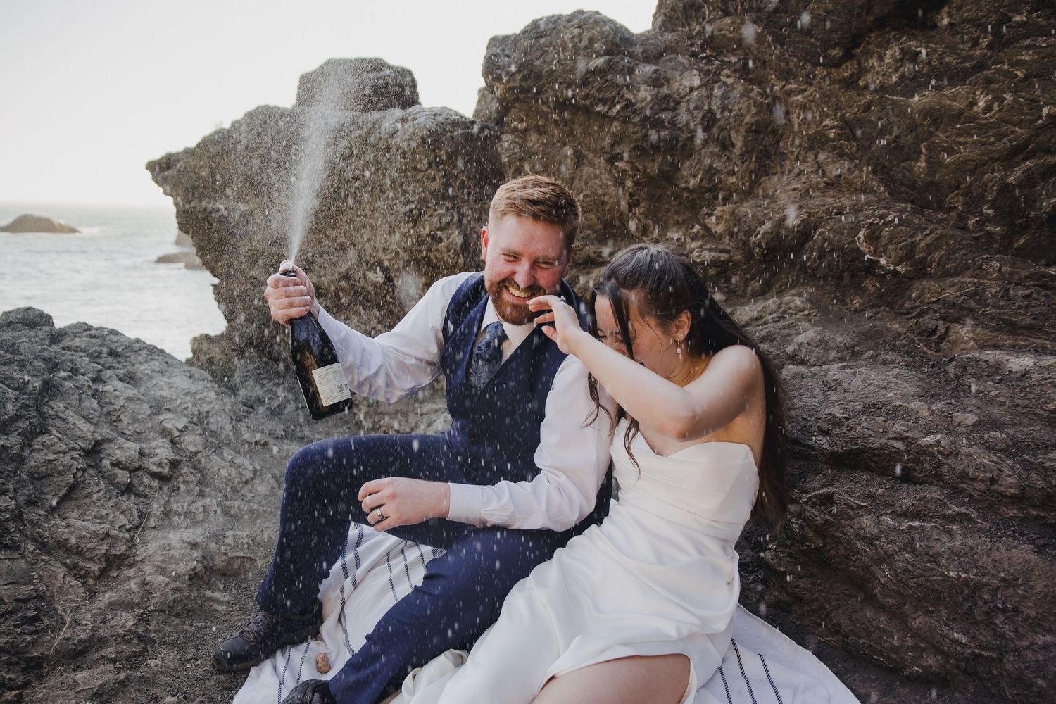 Couple in wedding attire celebrating on rocky beach, popping champagne bottle with ocean waves in background.