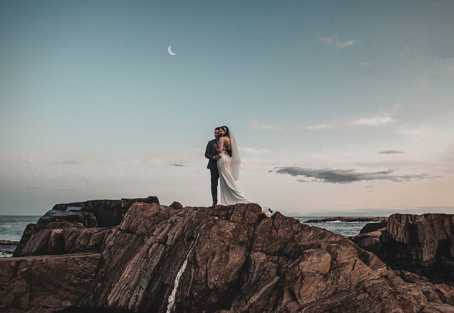 Wedding couple stands on rocky coastal cliffs at sunset with crescent moon in sky.