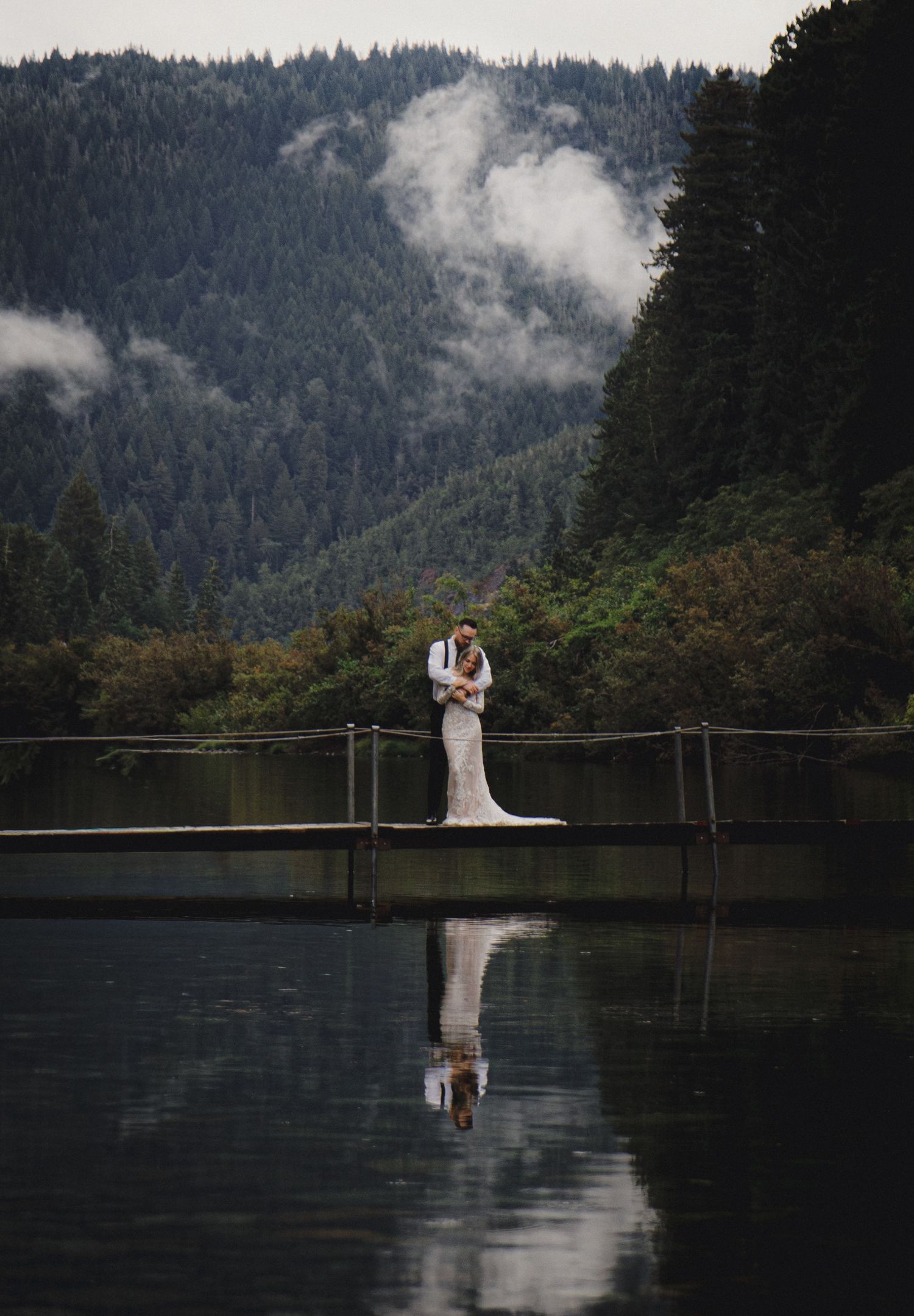 Bride and groom embrace on a dock over a still lake with misty mountain and forest backdrop, perfect reflection.