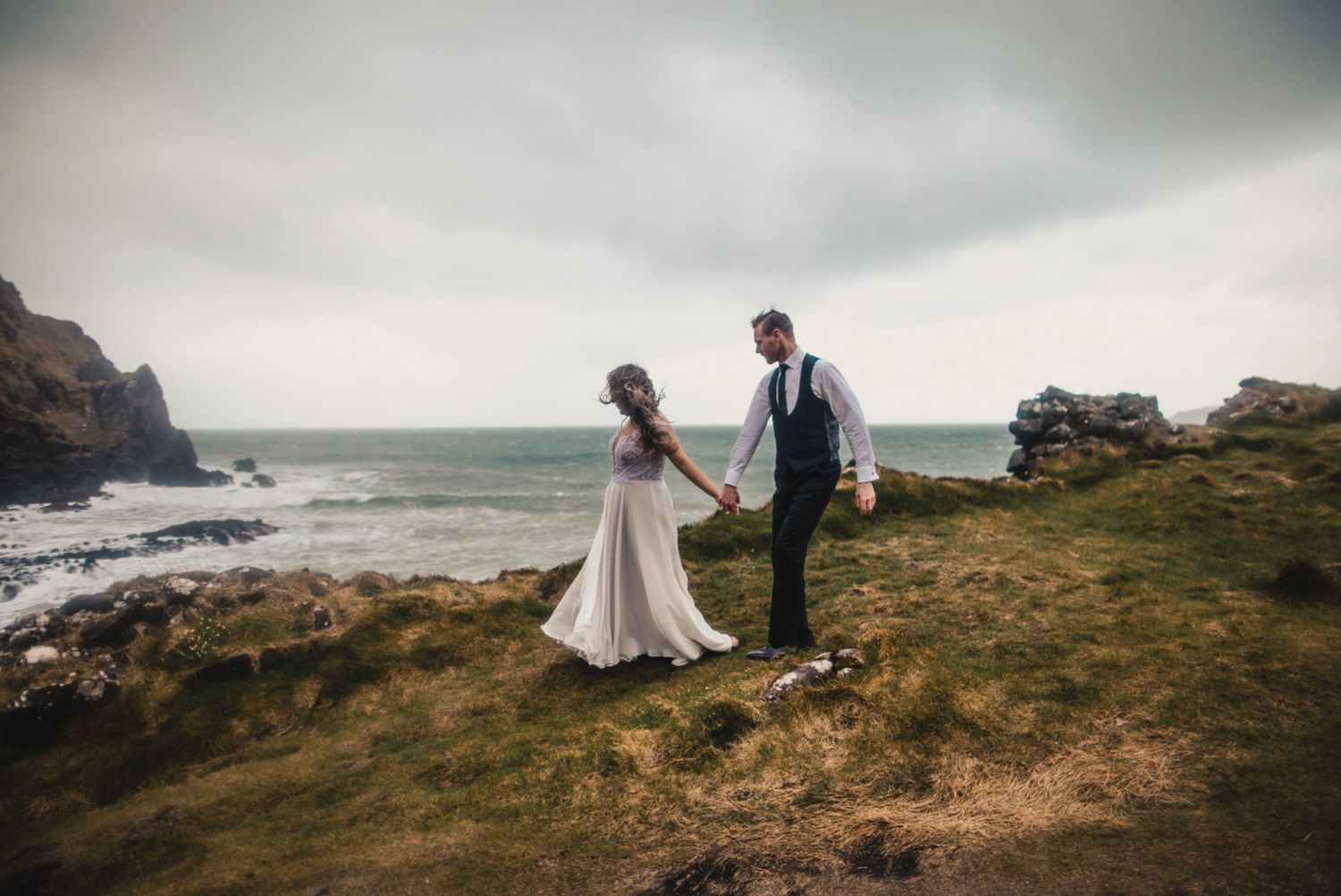 Couple in wedding attire poses on windswept coastal cliffs with ocean waves in background.