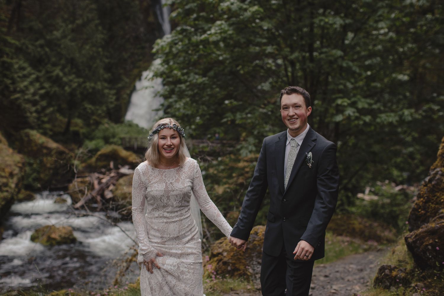 Wedding couple standing together near cascading waterfall in lush forest setting.