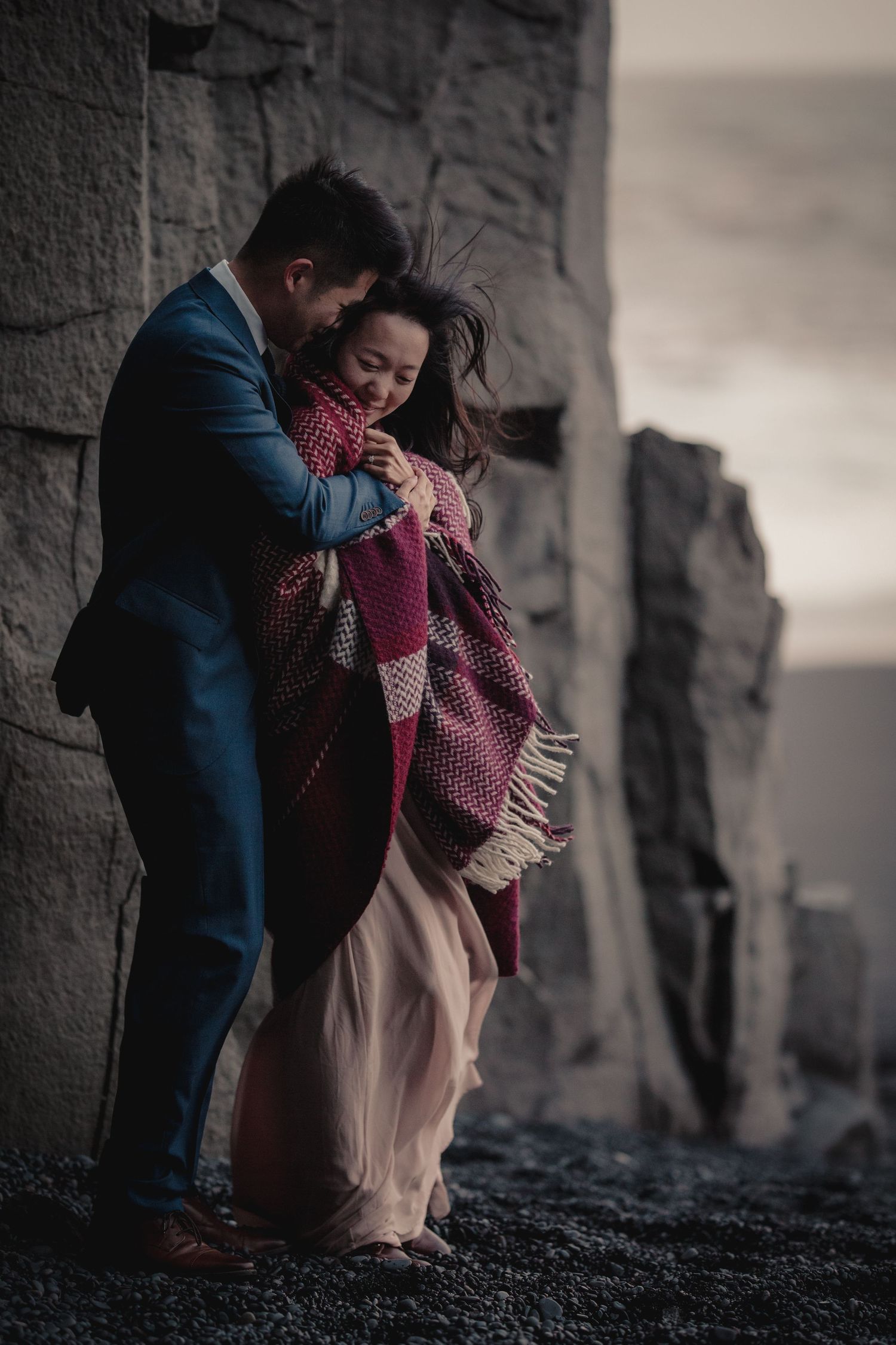 A romantic embrace against dramatic rocky cliffs with a couple wrapped in a cozy blanket at dusk.