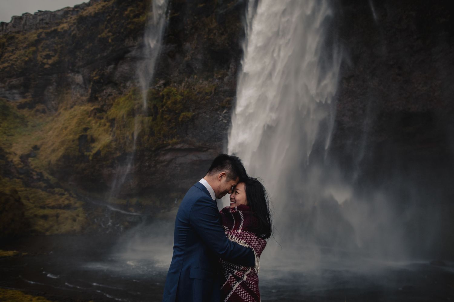 Romantic couple embracing near a towering waterfall against a dramatic rocky landscape.