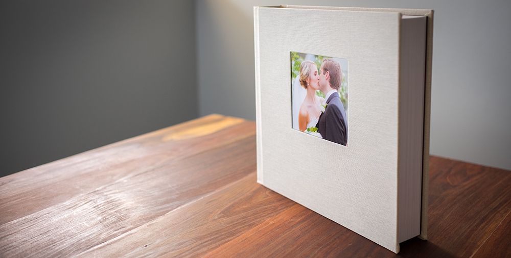 Elegant white linen wedding photo album with couple portrait on cover, displayed on a wooden table.