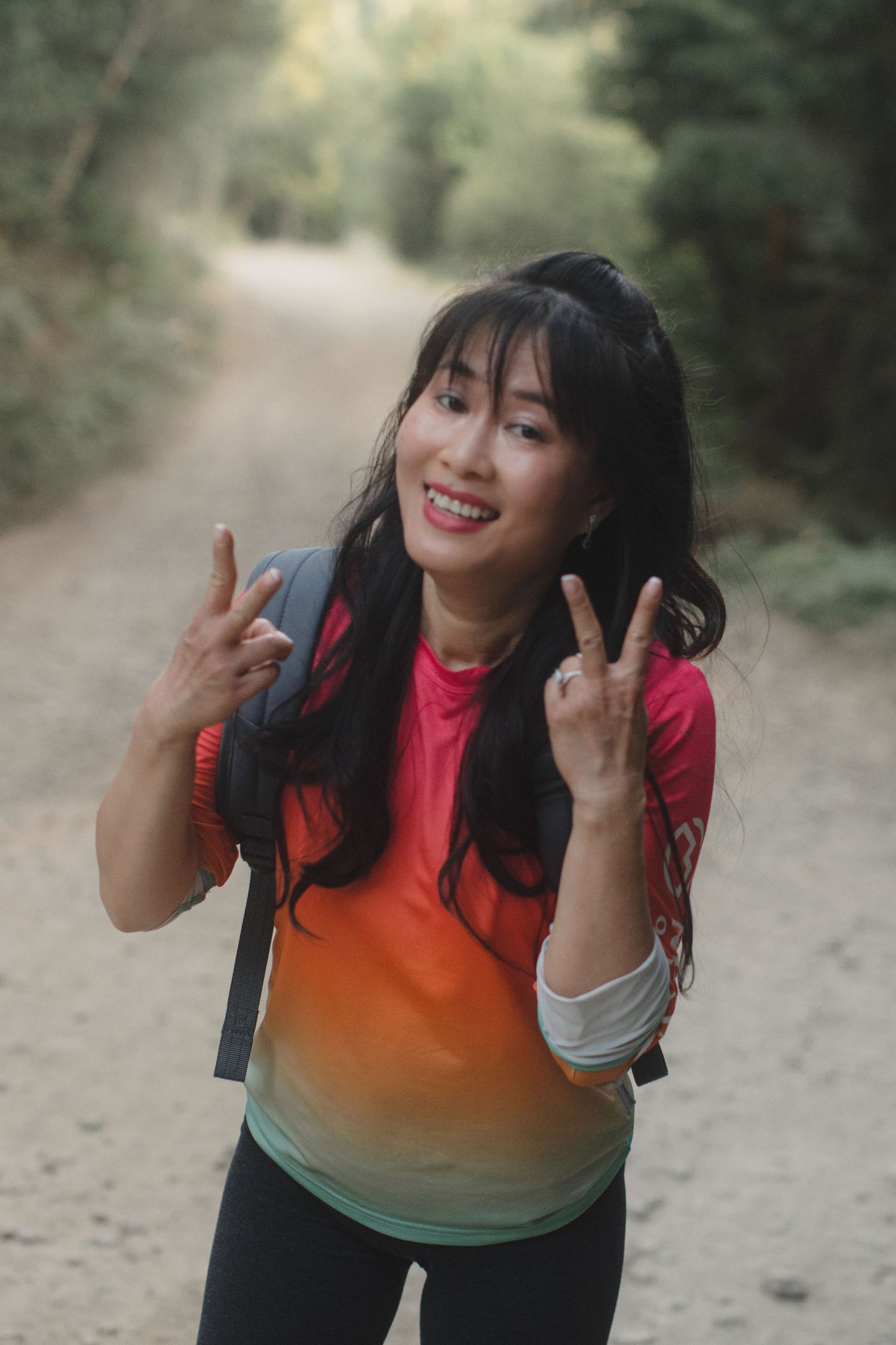 Smiling Asian woman making peace signs on a forest trail with a backpack.