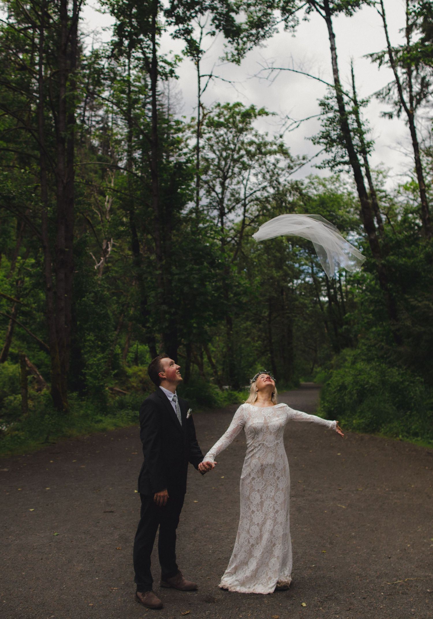 A couple shares a playful moment on a forest path with the bride's veil catching the wind.