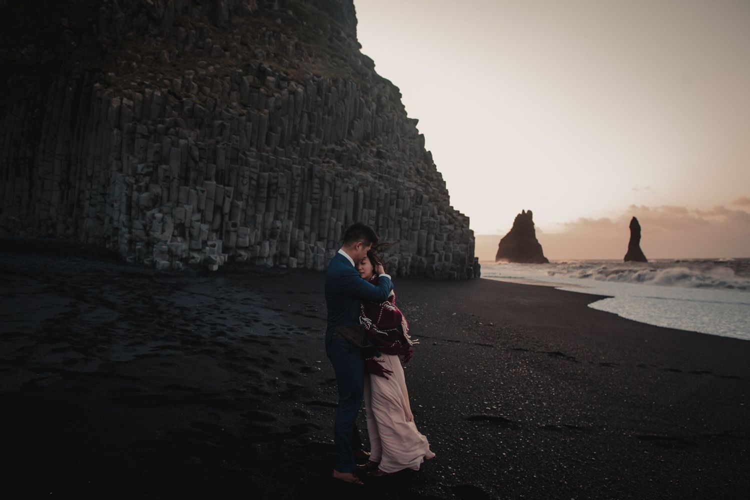 A romantic scene unfolds on a black sand beach with dramatic basalt cliffs and sea stacks at sunset.