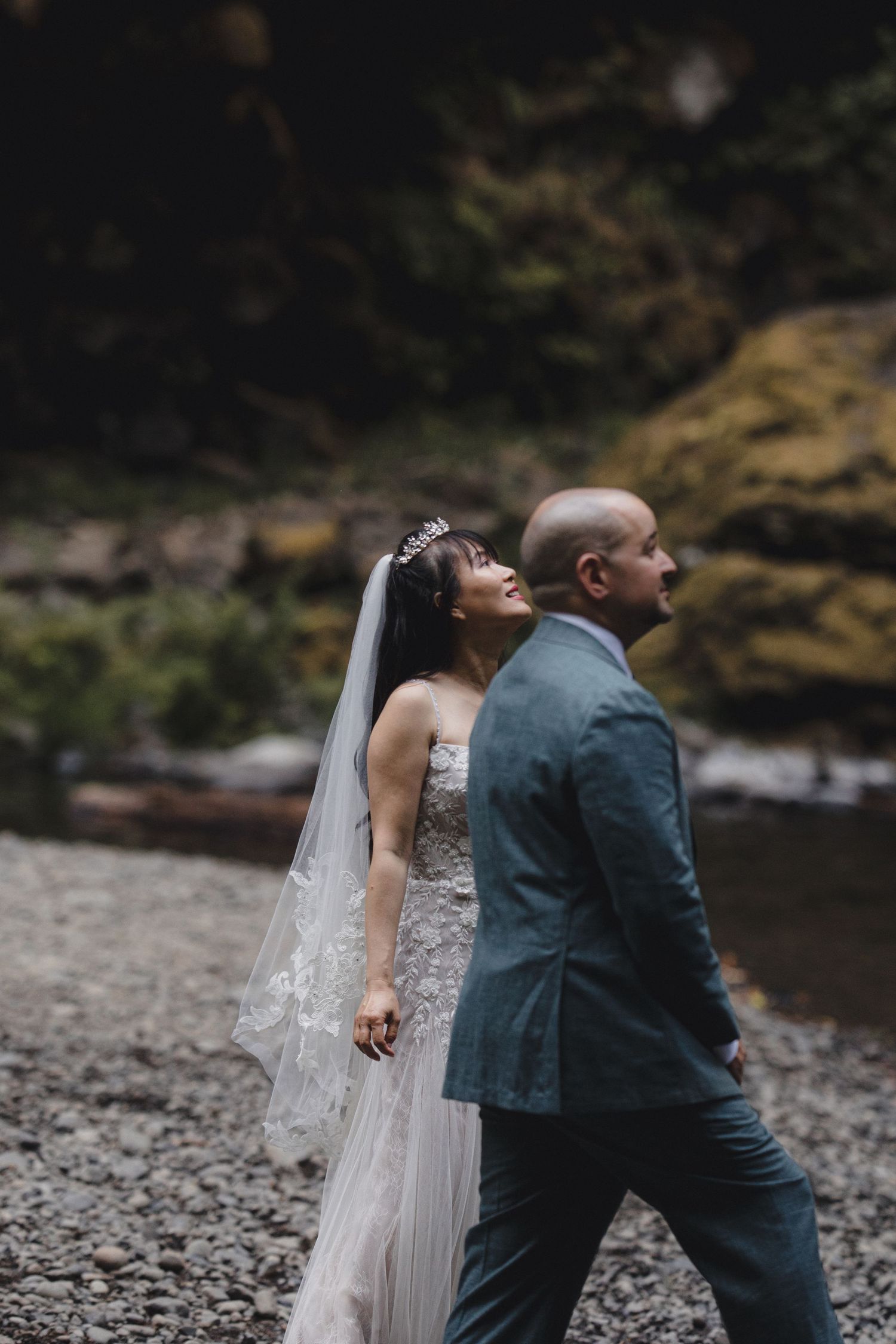 Bride and groom walking along rocky riverbank in lush forest during outdoor wedding ceremony.