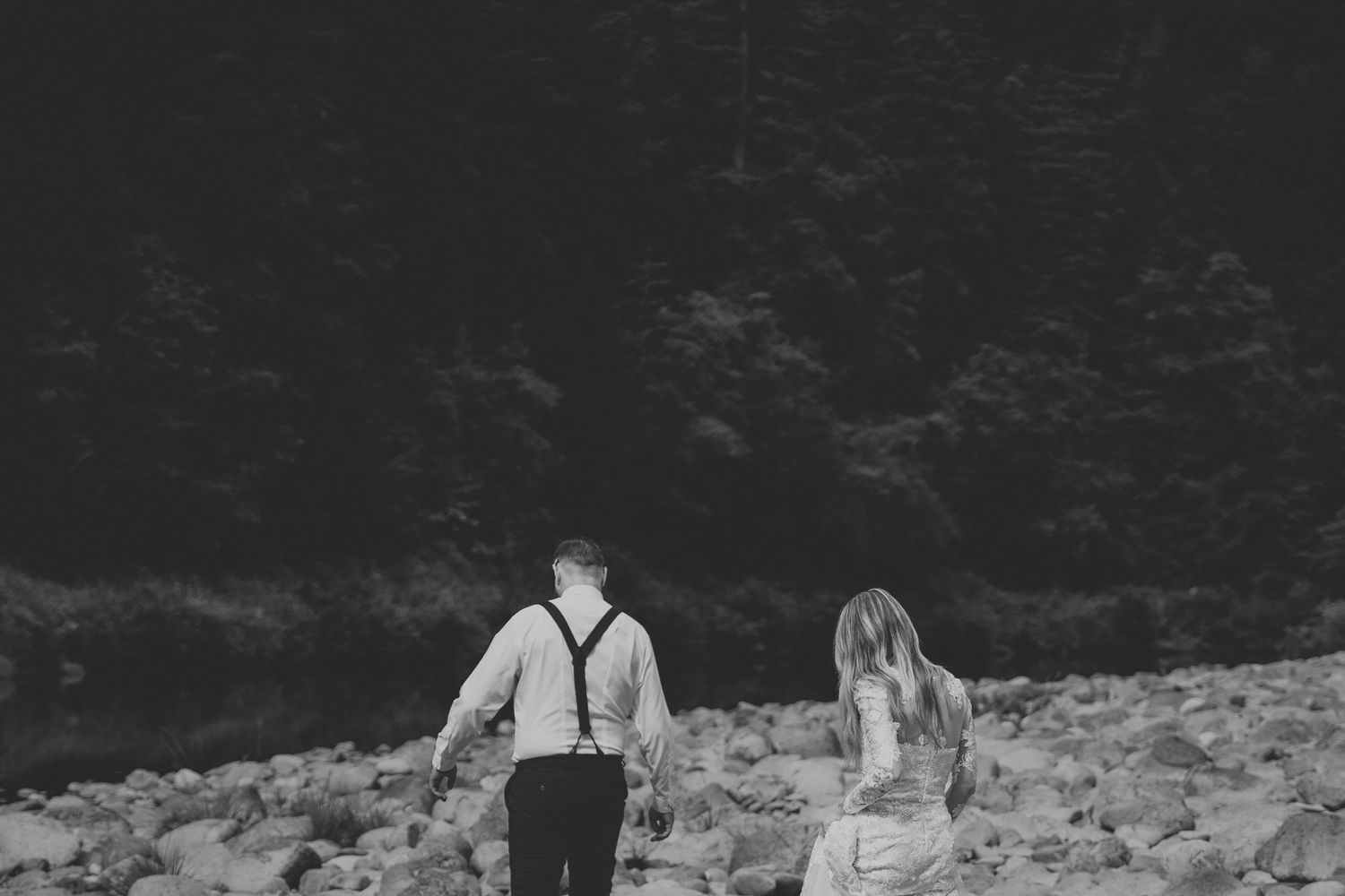 Black and white photo of a couple holding hands walking on rocky terrain with dark forest trees in the background.