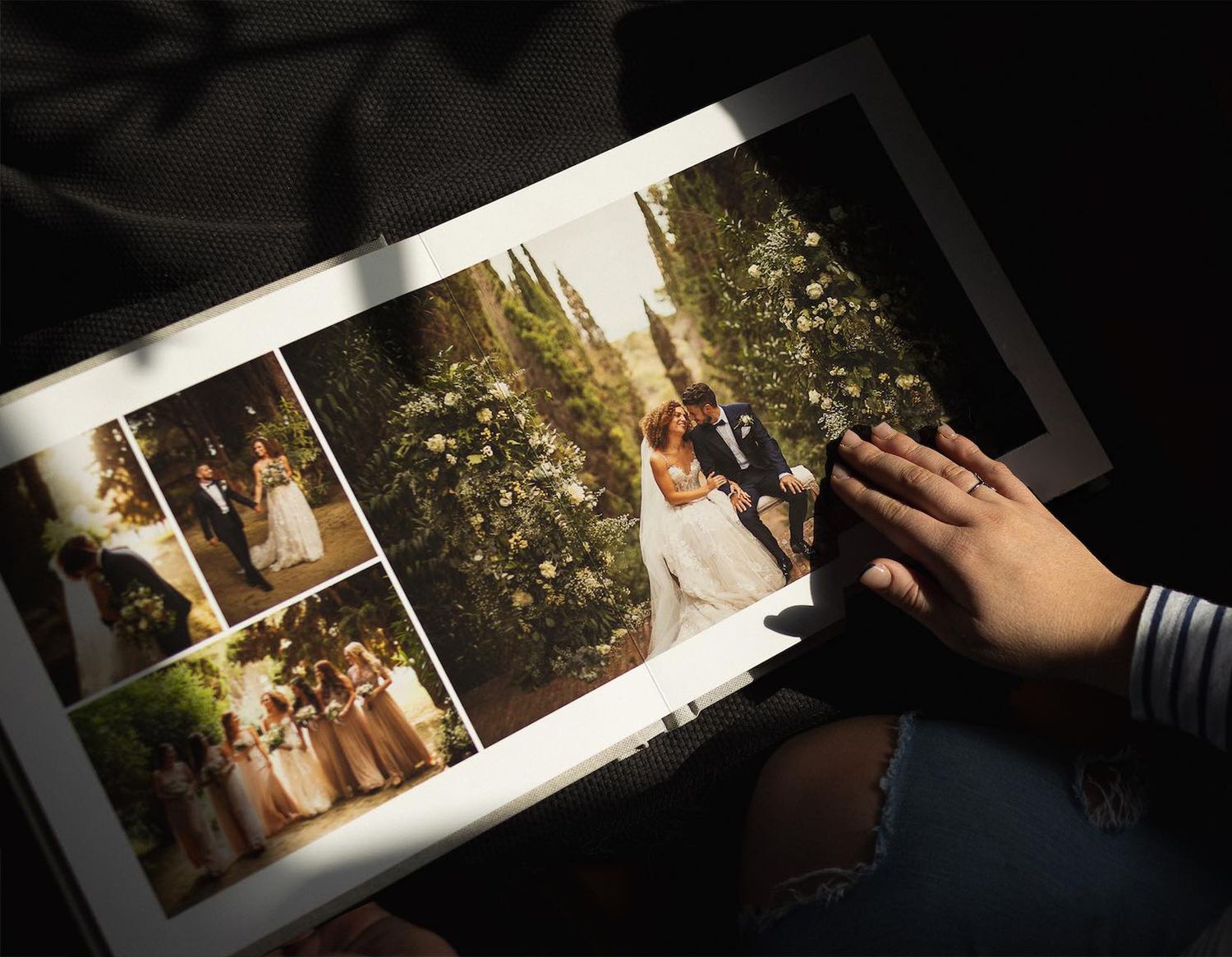 Person holding an open wedding photo album featuring bride and groom portraits surrounded by lush greenery.