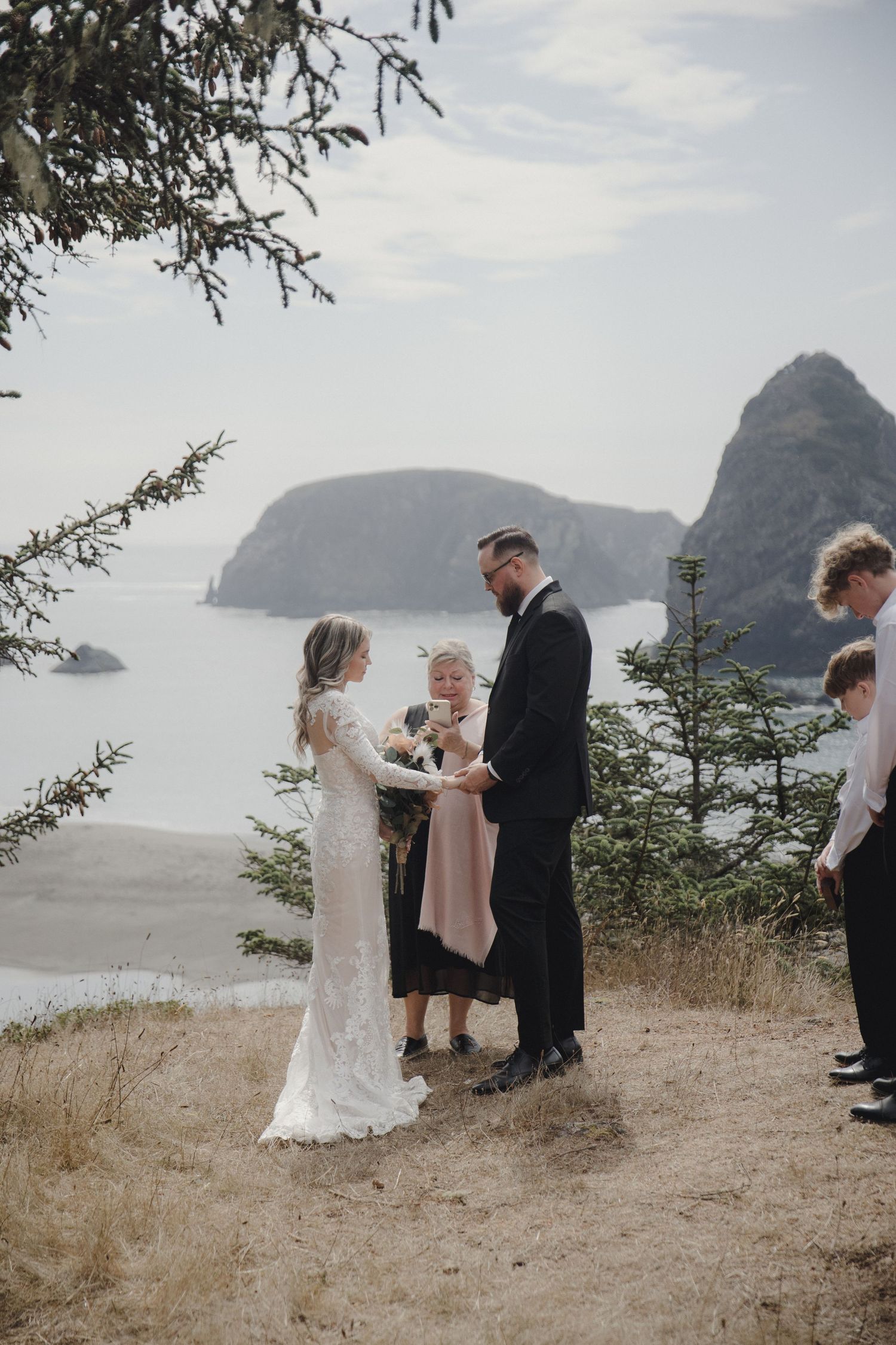 Couple exchanges vows at outdoor coastal wedding ceremony with dramatic rock formations and ocean views in background.