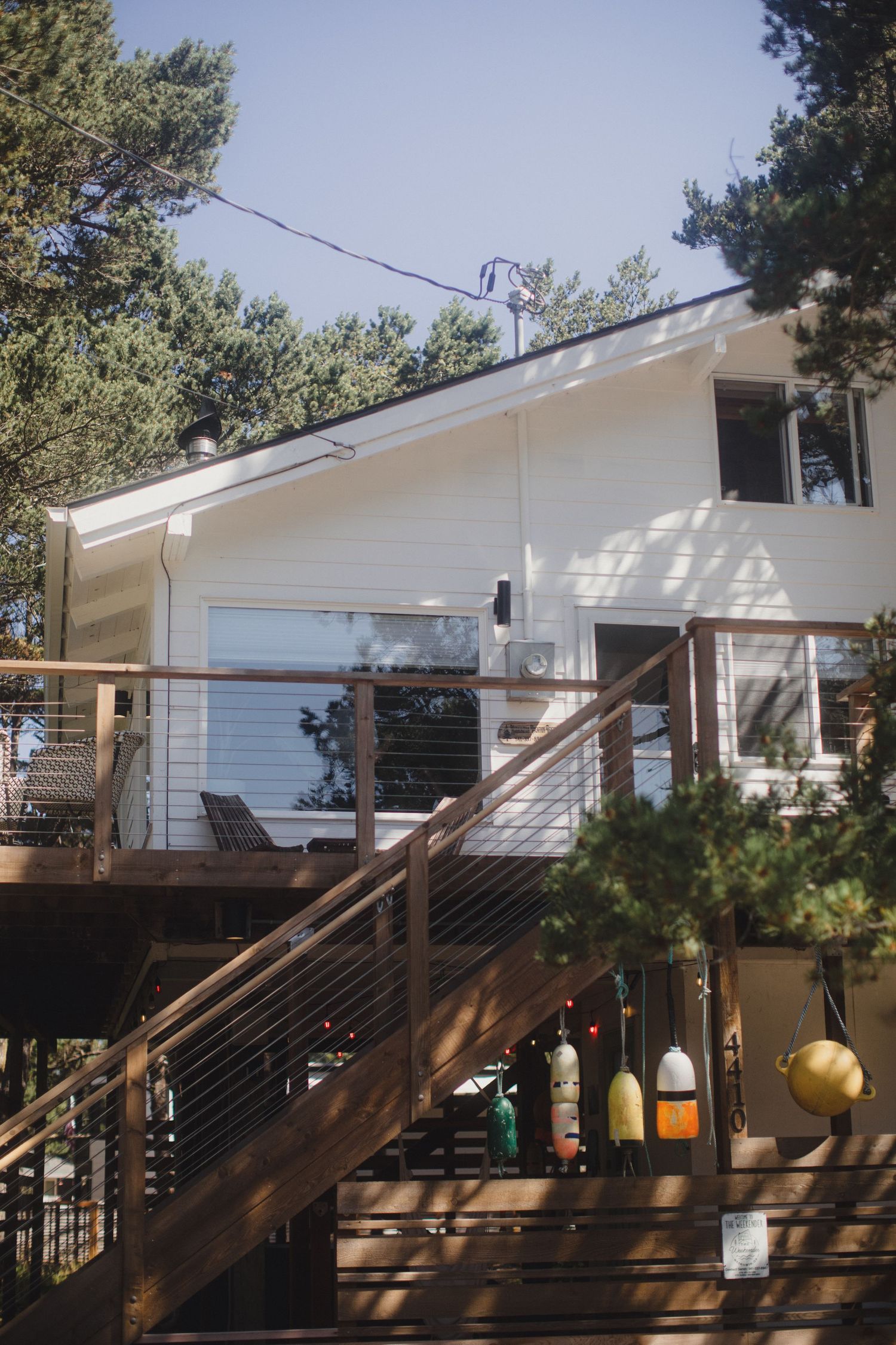Elevated beach house with wooden deck, cable railings, and stairs surrounded by tall pine trees.