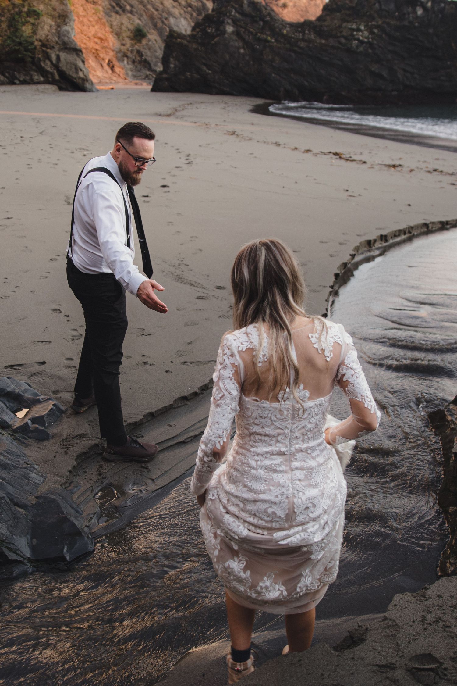 Groom reaches out his hand to bride in a lace wedding dress on a secluded sandy beach at dusk.