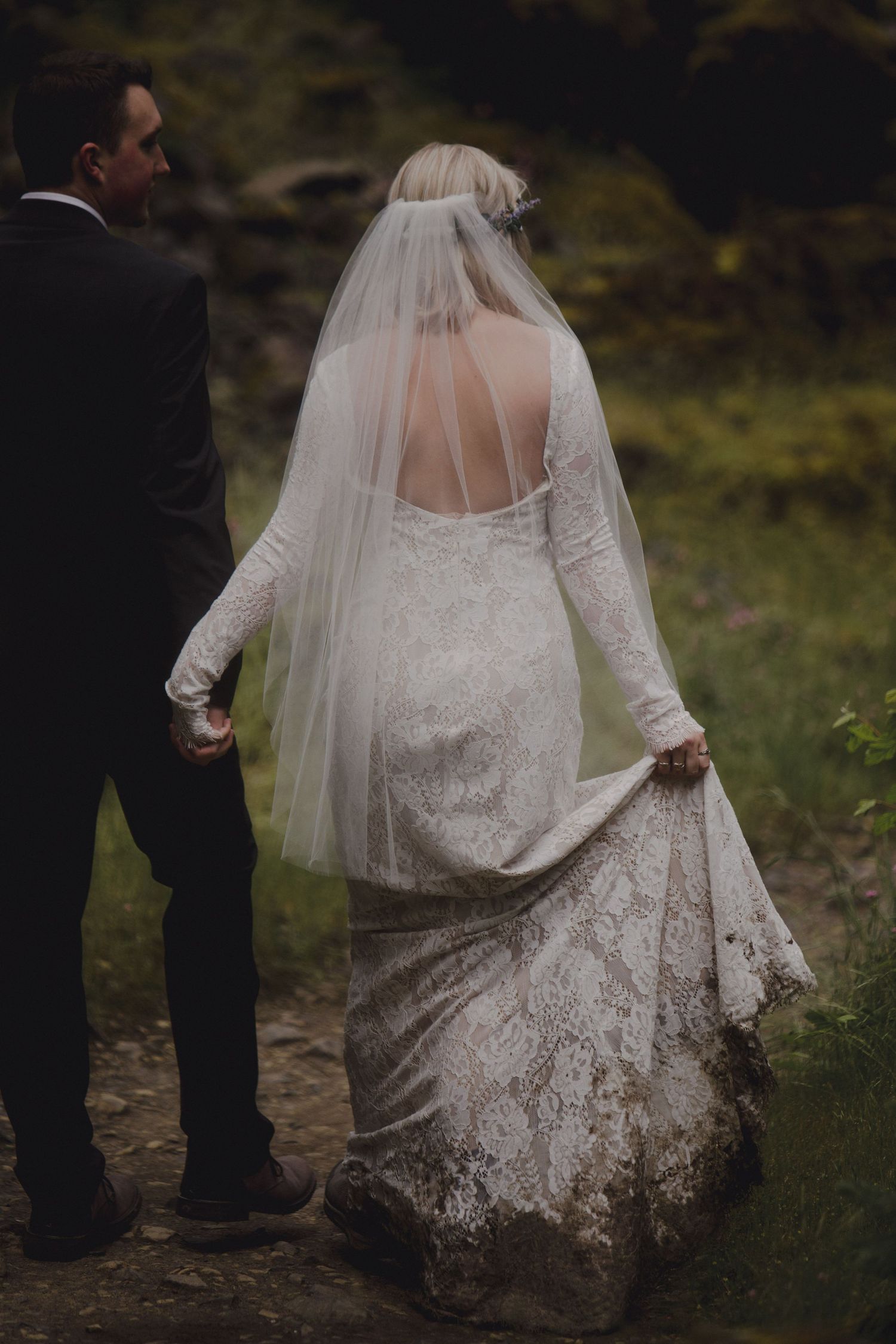 Back view of a bride in lace dress and veil walking with groom on a dirt path.