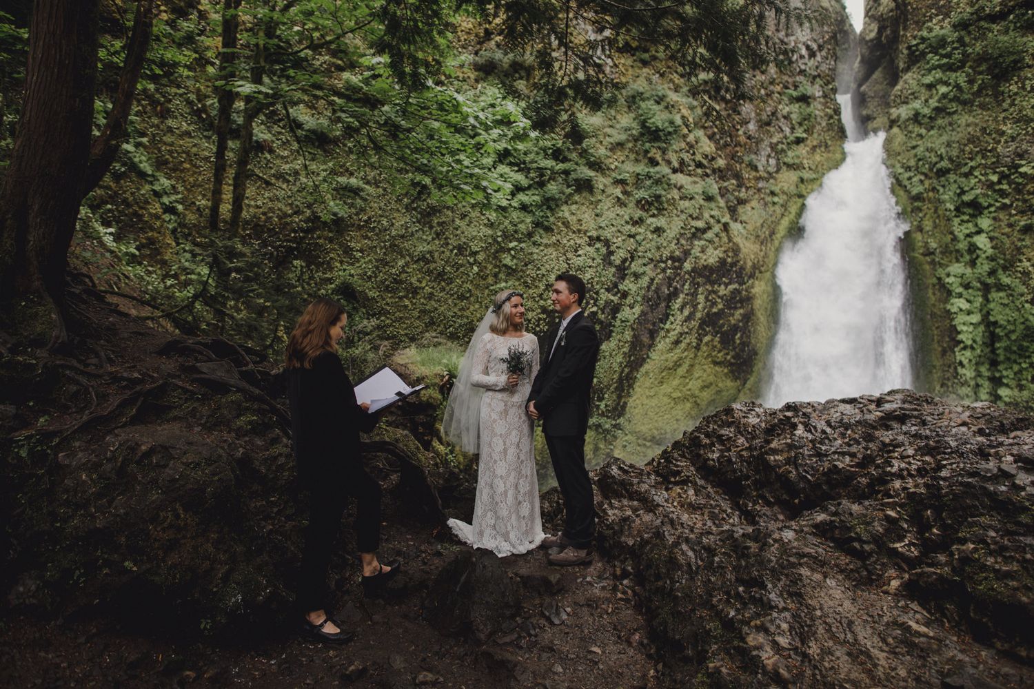 An elopement ceremony takes place beside a cascading waterfall in a lush forest setting.