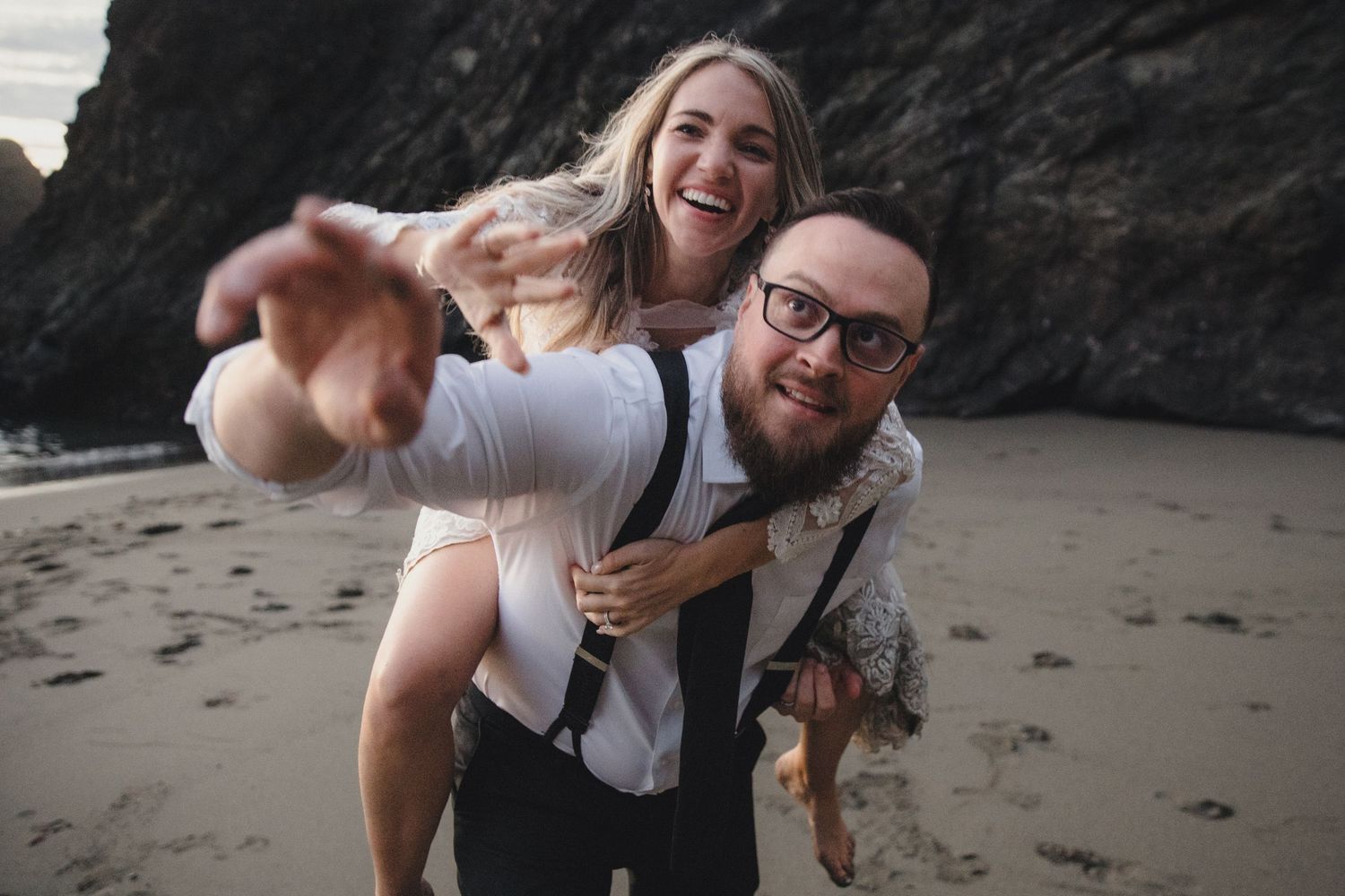 A joyful couple on a beach, man giving woman a piggyback ride, both laughing and pointing, wearing semi-formal attire.