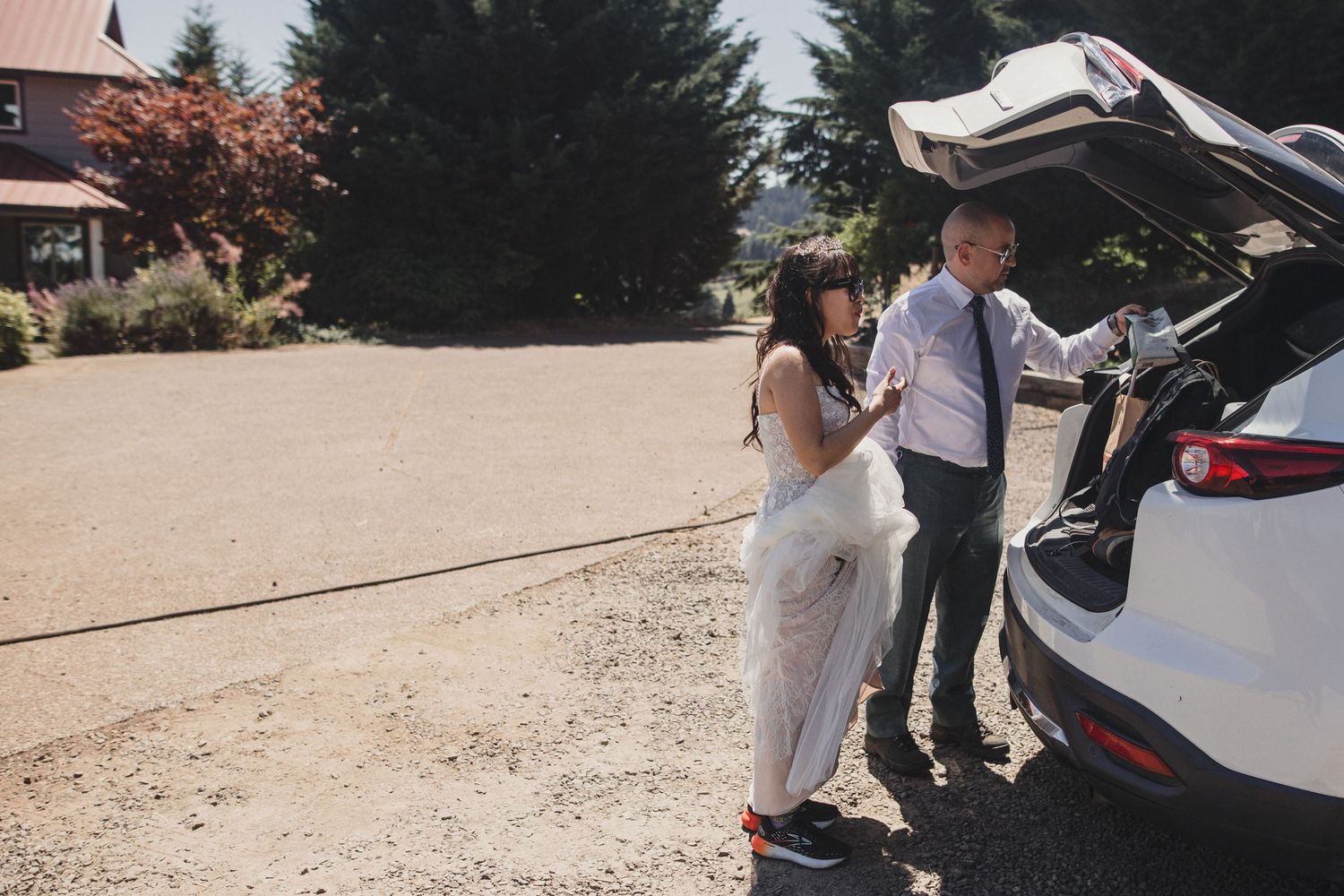 Bride in white dress and groom in suit unloading car trunk outdoors on sunny wedding day.