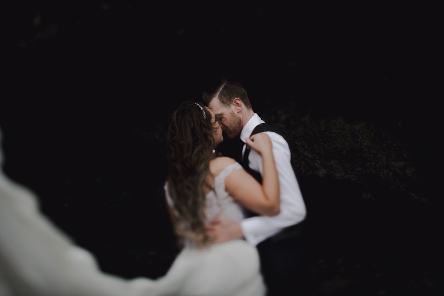 A romantic wedding couple embraces in a dramatic dark setting with ethereal lighting.
