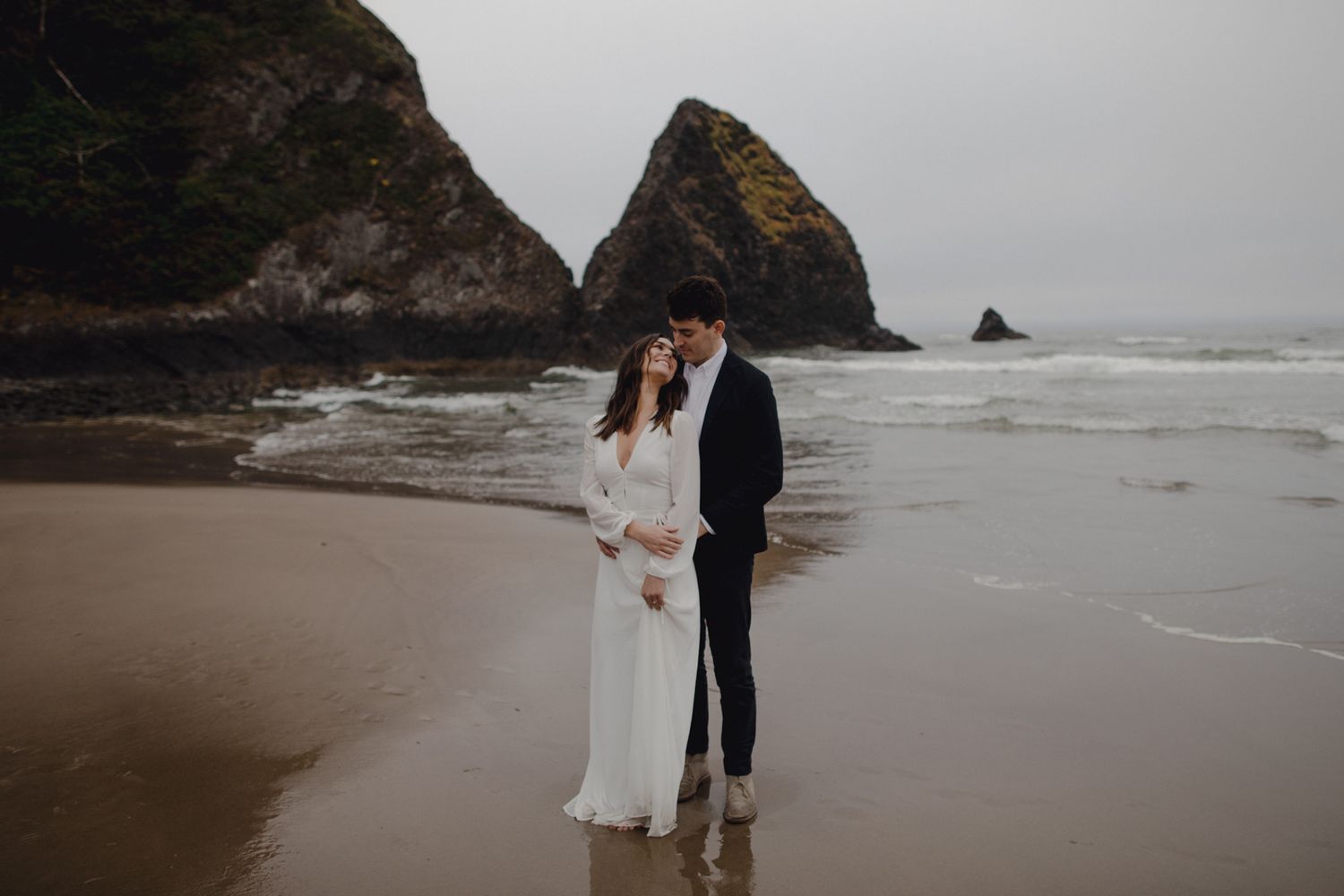 Couple in wedding attire embracing on a misty Oregon beach with dramatic sea stacks in the background.