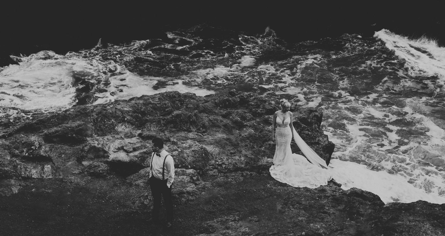 A dramatic black and white wedding photo captured on a rugged coastal cliff with crashing waves below.