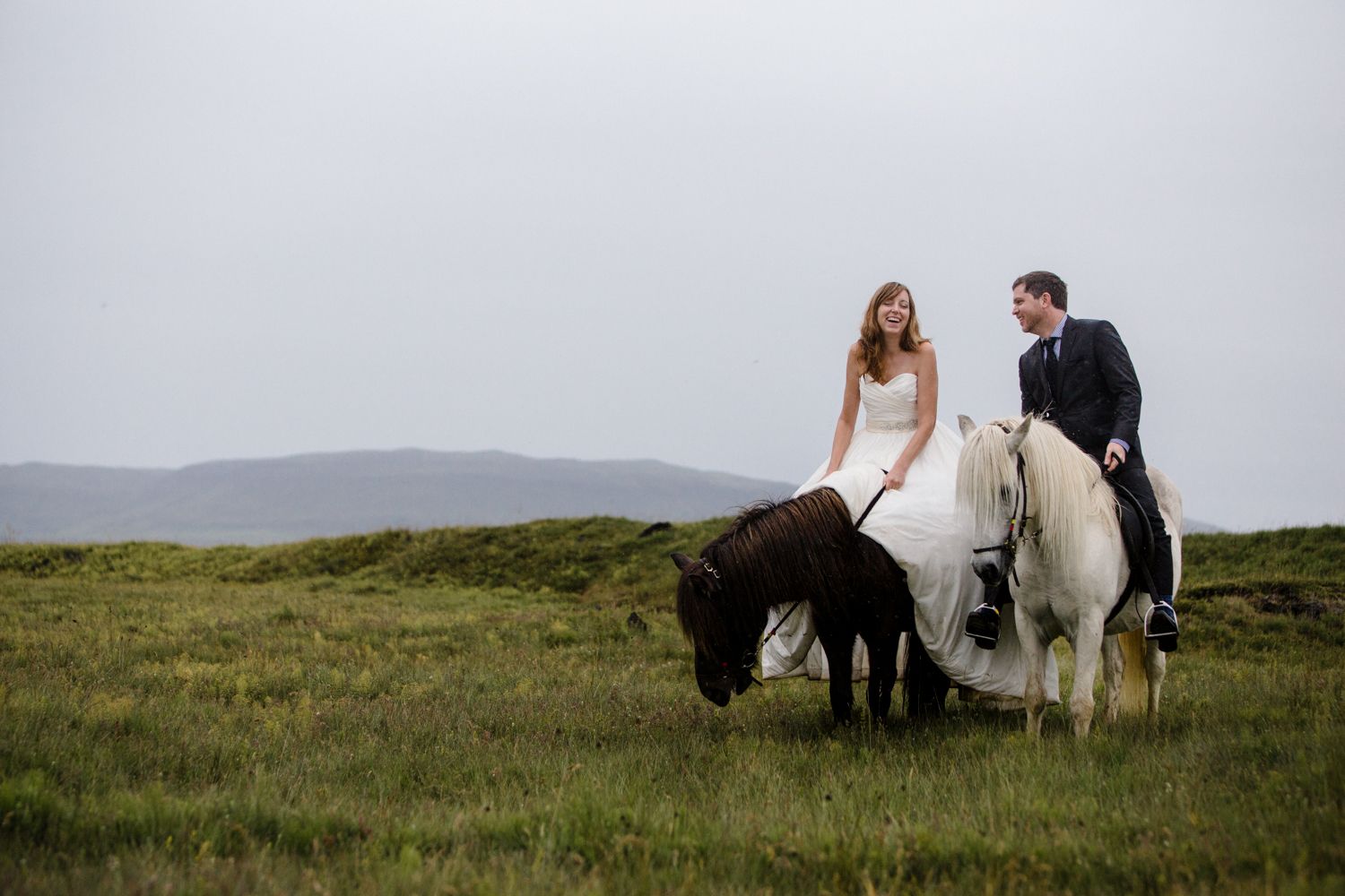Bride and groom in wedding attire riding horses across a lush green field with misty hills in the background.