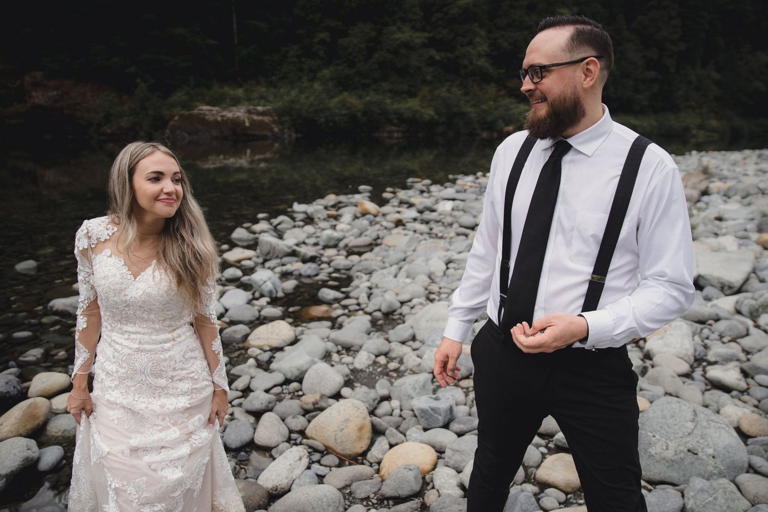 Bride in lace gown and groom in white shirt with suspenders share a joyful moment on a rocky riverbank.