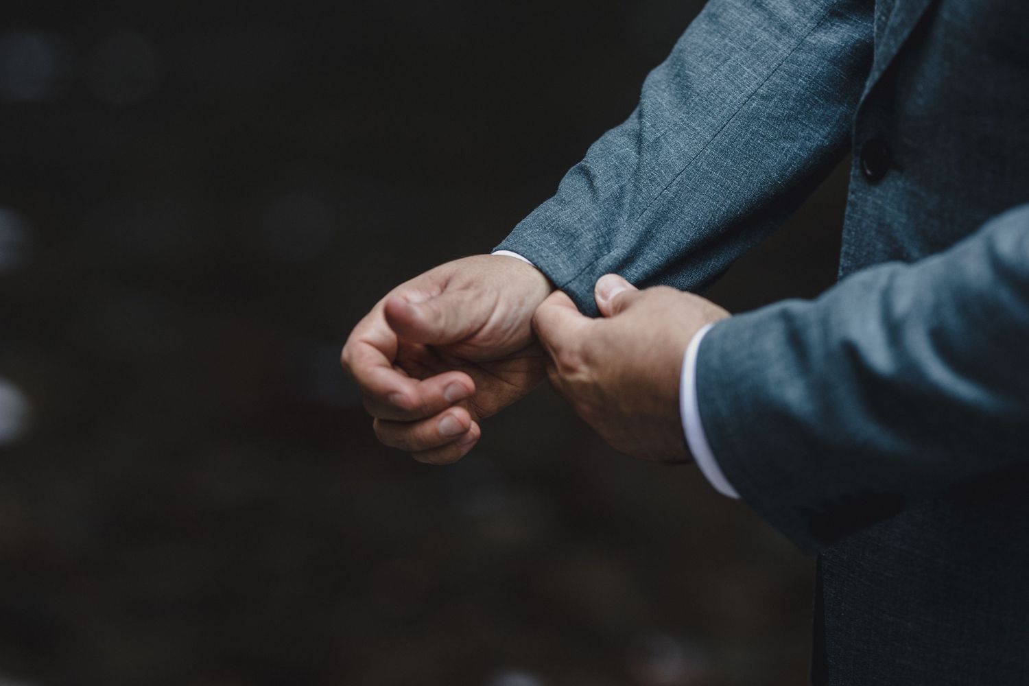 Groom in grey suit adjusting his cufflinks against a dark blurred background.