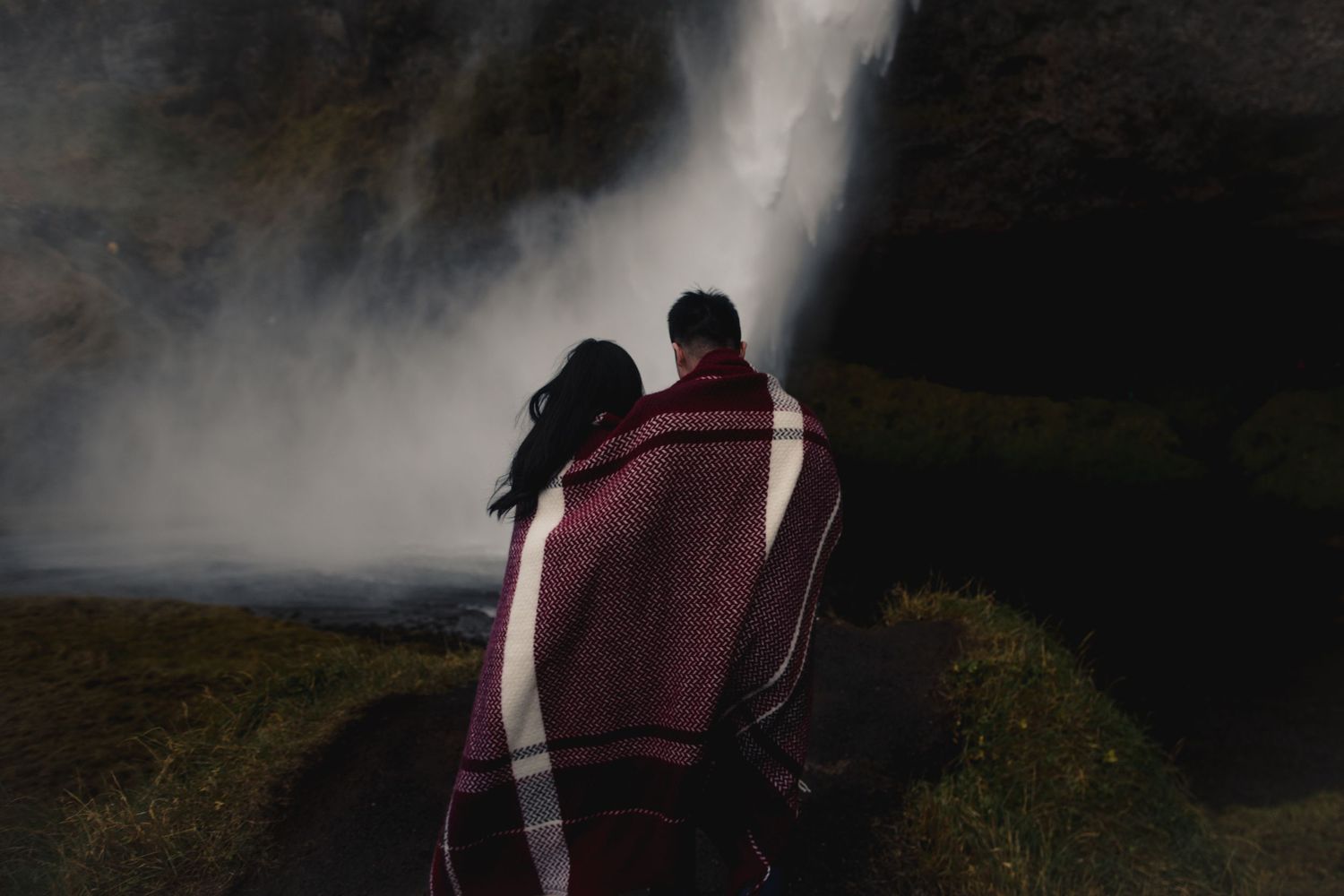 A couple wrapped in a plaid blanket stands before a majestic waterfall in moody lighting.