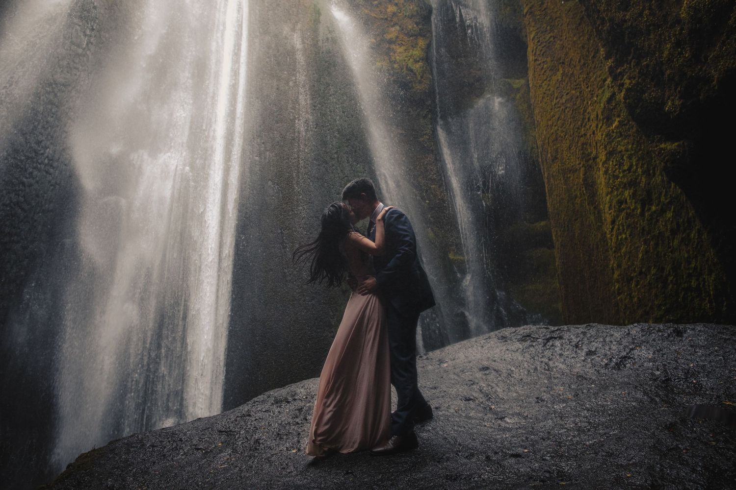 Couple in formal attire embracing under dramatic waterfall spray.