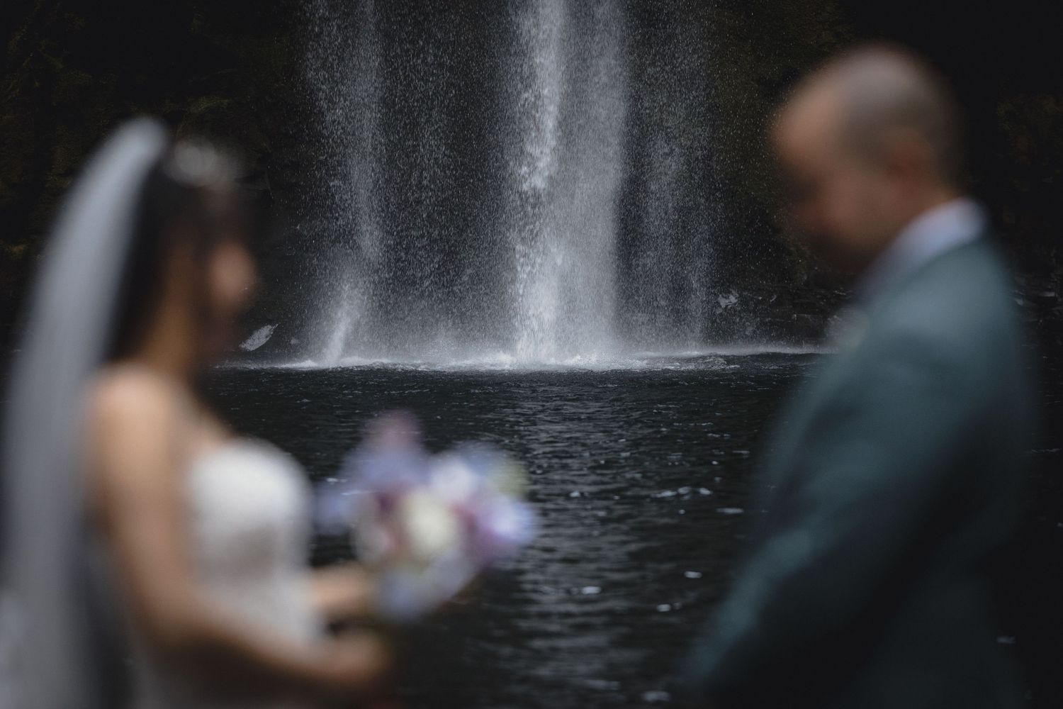 Blurred bride and groom standing before a dramatic waterfall holding a floral bouquet.