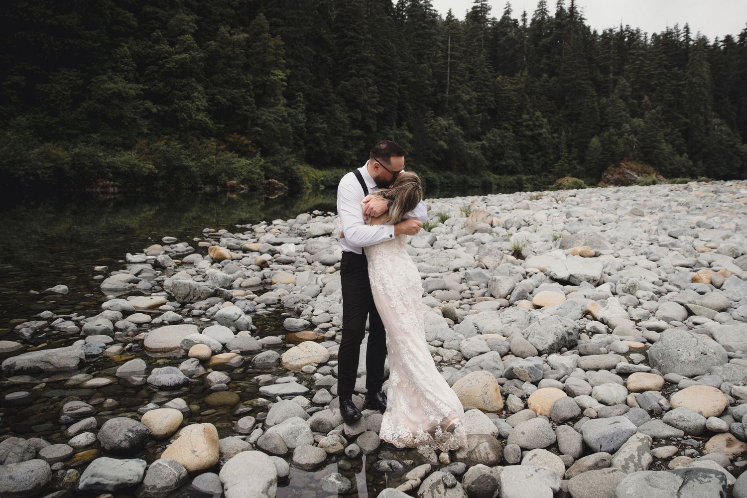 Bride and groom embrace on rocky riverbank surrounded by lush evergreen forest during elopement.