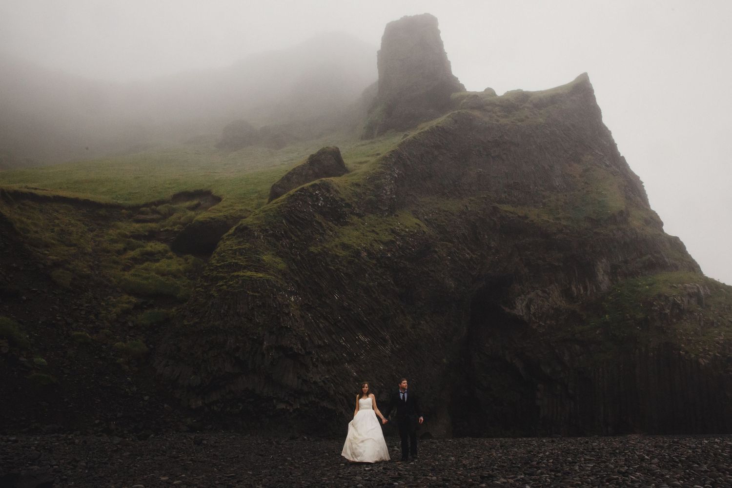 Bride and groom stand on black sand beach before dramatic mossy cliffs in misty Iceland landscape.