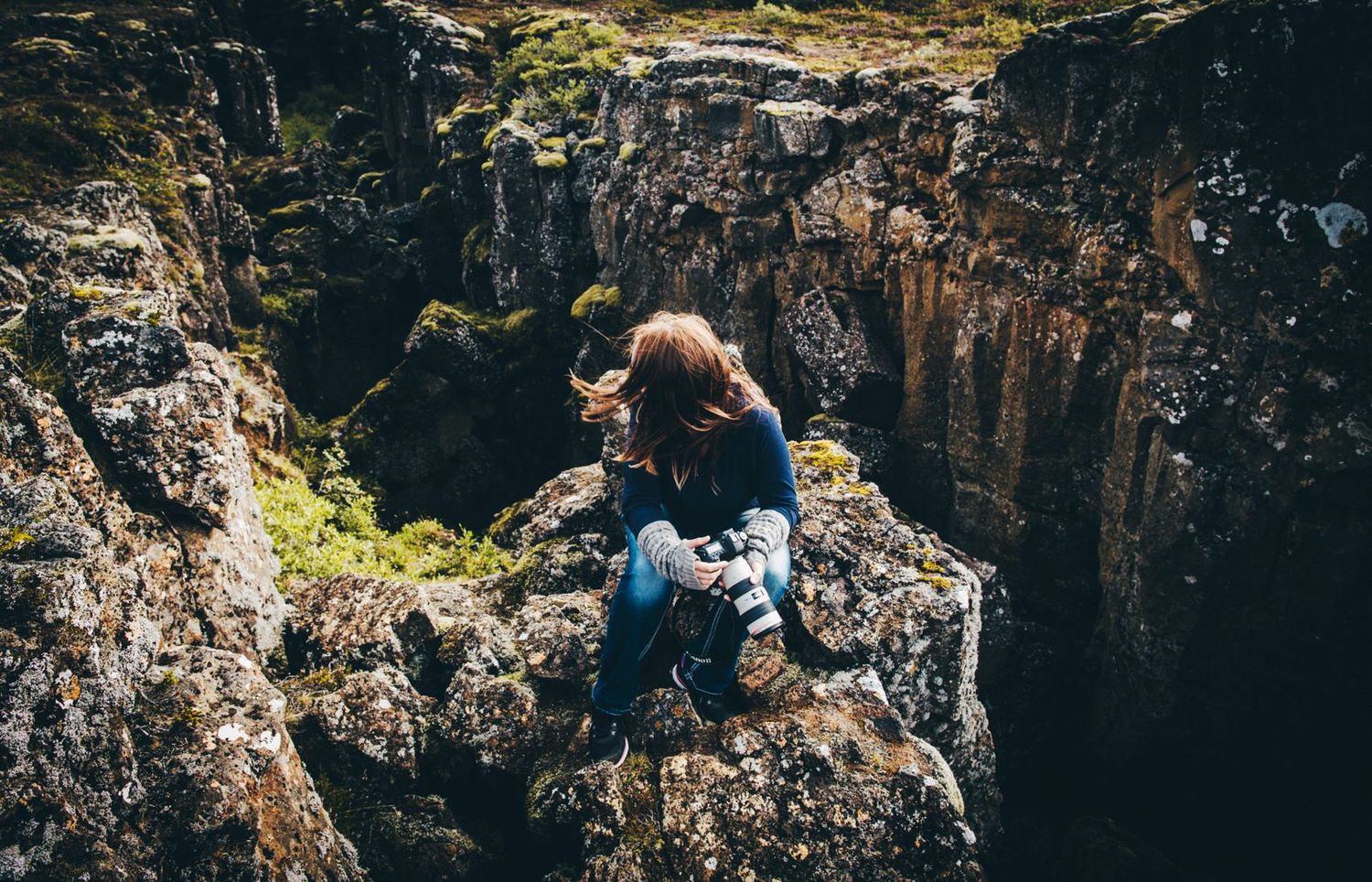 A woman sits on rocky cliffs edge holding a camera, surrounded by mossy boulders and a dramatic canyon below.