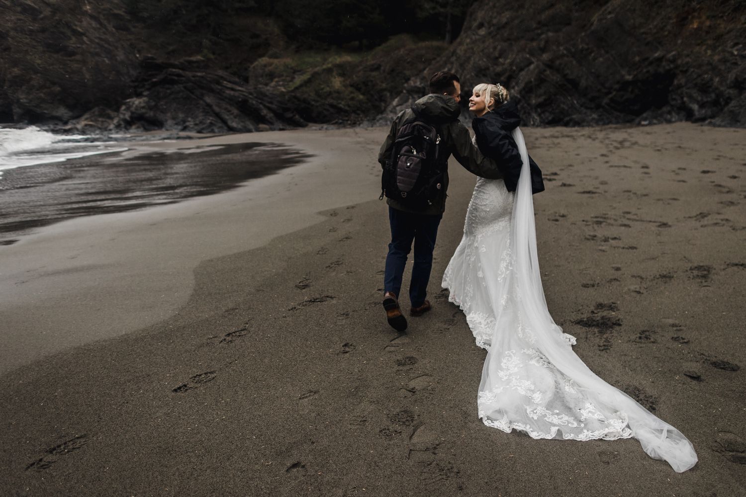 A couple walks along a moody black sand beach with flowing white wedding dress.
