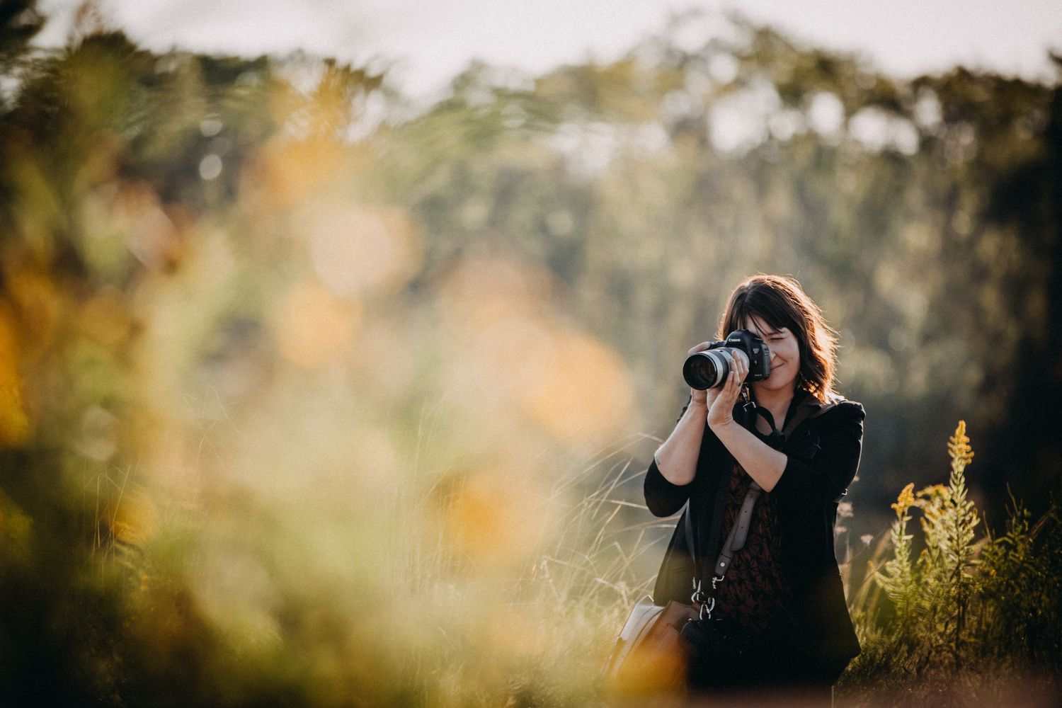 A woman photographer captures nature shots outdoors using a DSLR camera with a telephoto lens in a sunlit field.