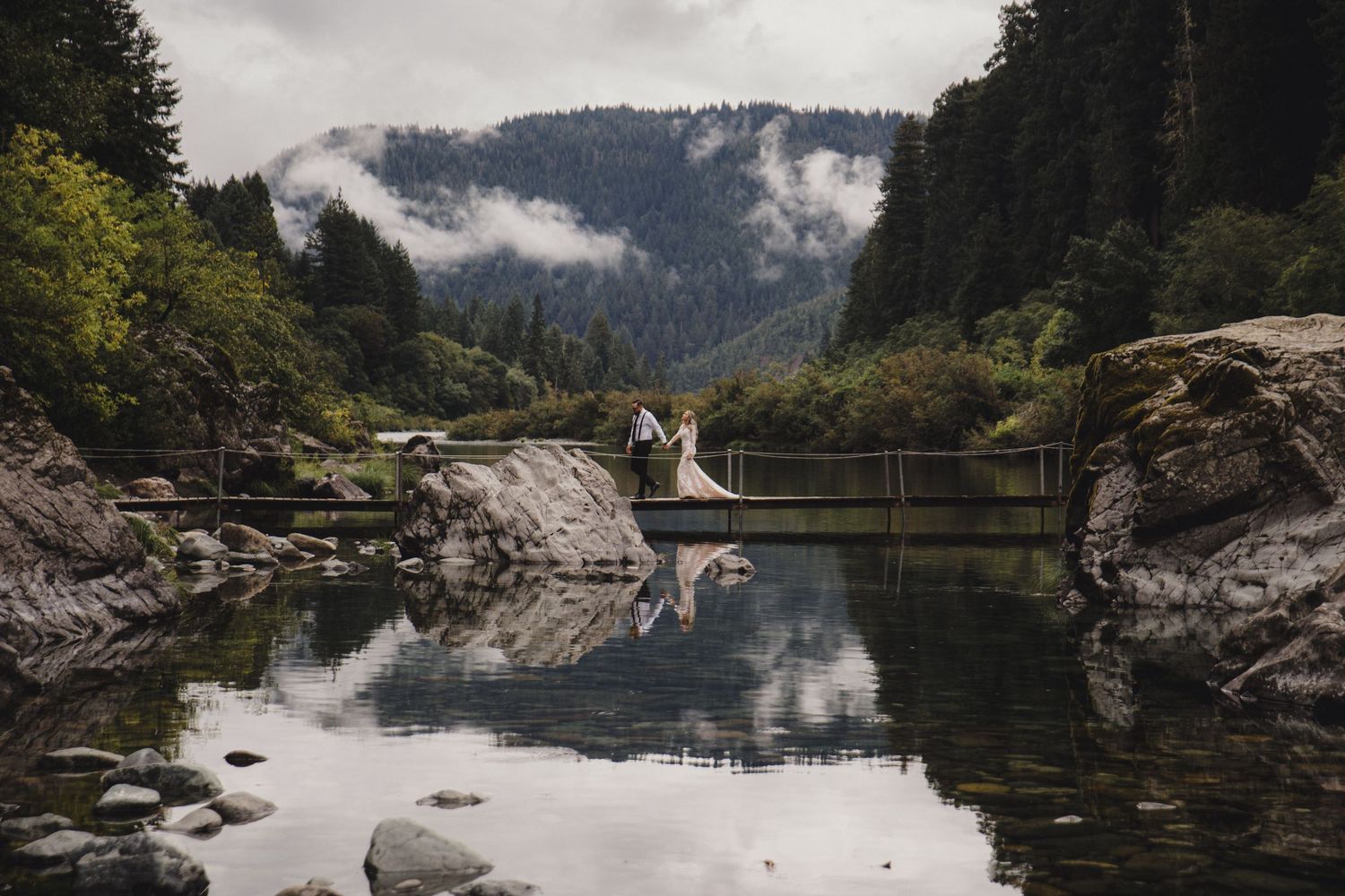 Bride and groom share a romantic moment on a suspension bridge reflecting in calm water surrounded by misty forested mountains.