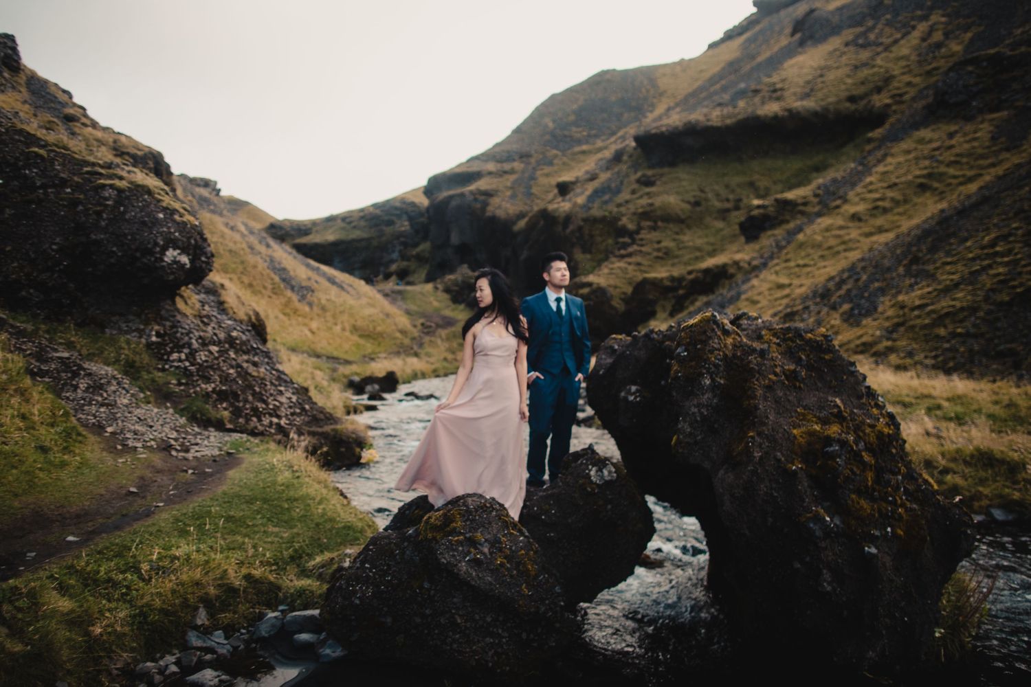 A couple stands on rocky cliffs overlooking a winding river valley in moody Icelandic landscape.