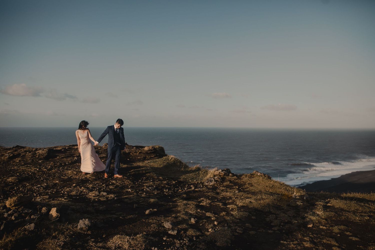 A couple walks along dramatic coastal cliffs overlooking ocean waves at dusk.