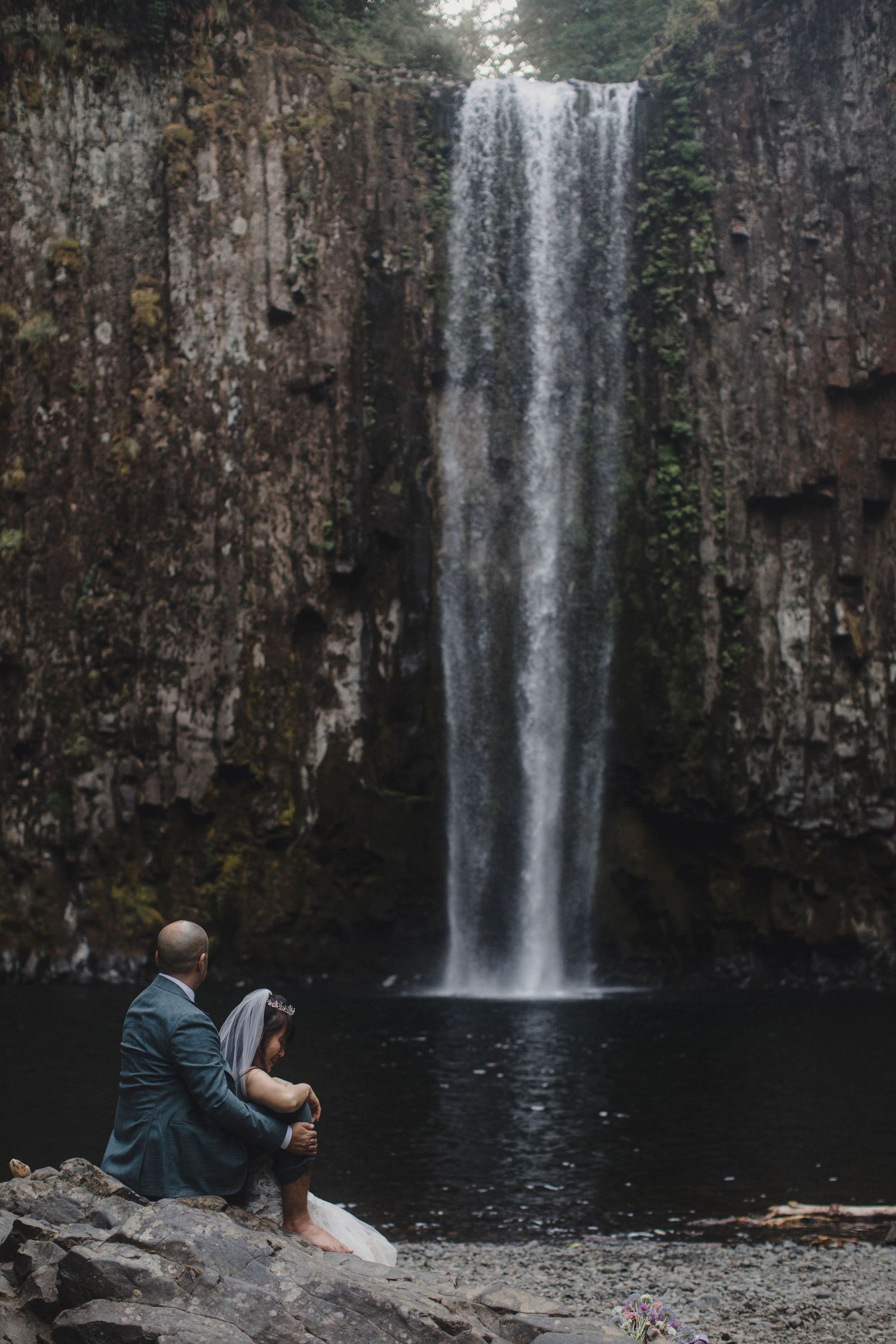 Bride and groom sitting together on rocks before a tall cascading waterfall in nature.