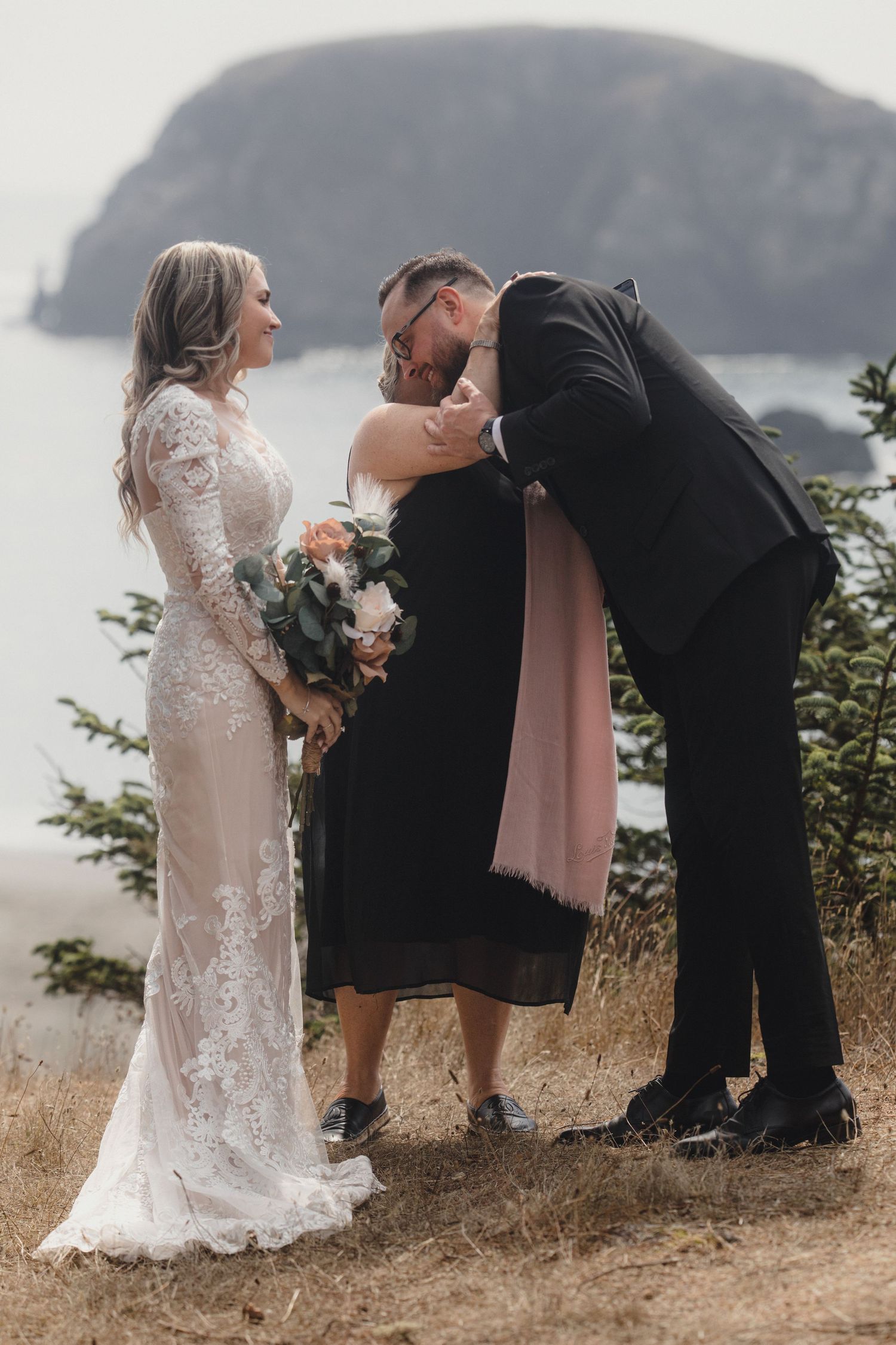 Groom kissing wedding officiant on the head during outdoor coastal cliff ceremony with ocean views.