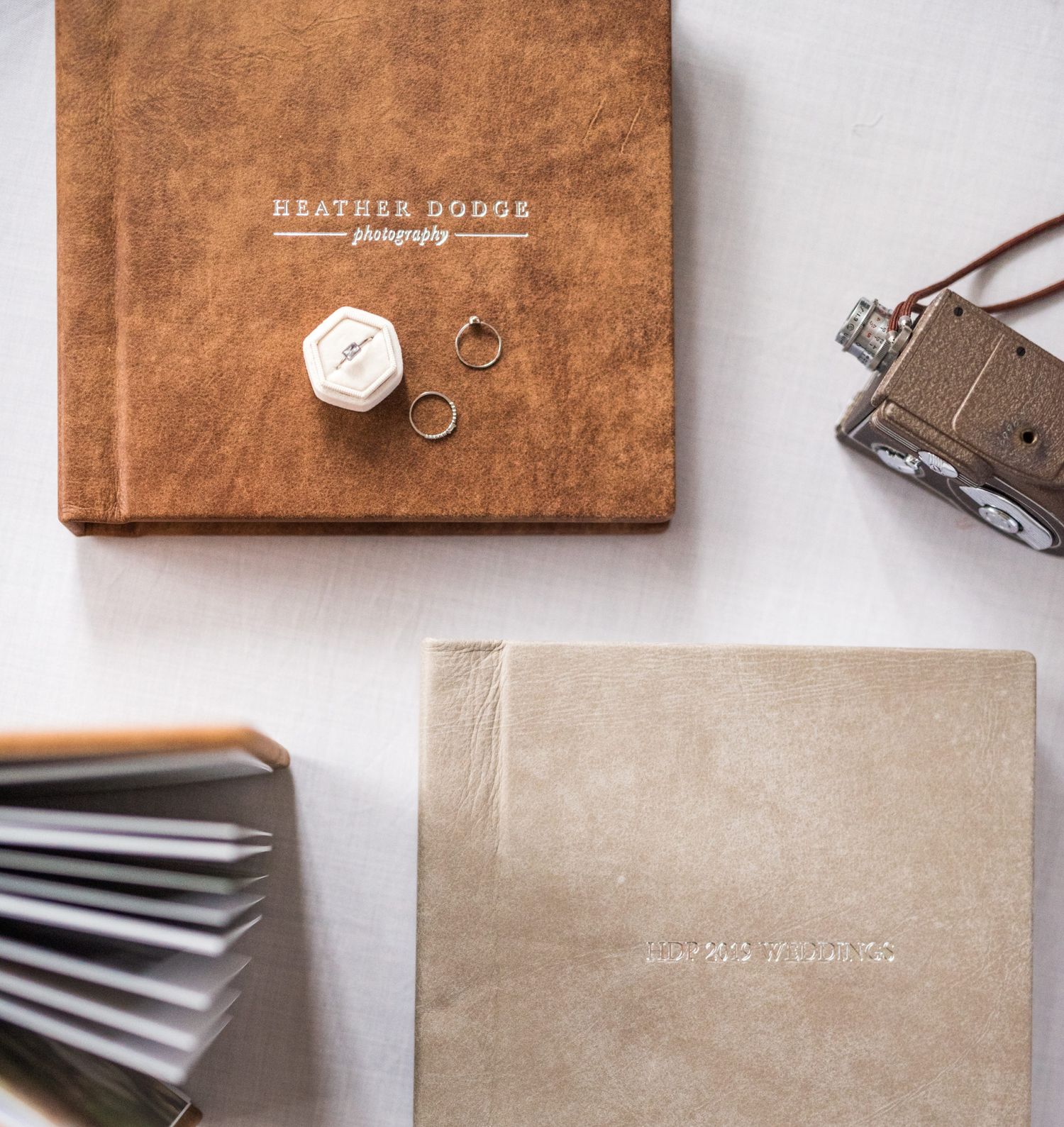 Wedding rings, vintage camera, and leather photo albums by Heather Dodge Photography displayed on a white surface.