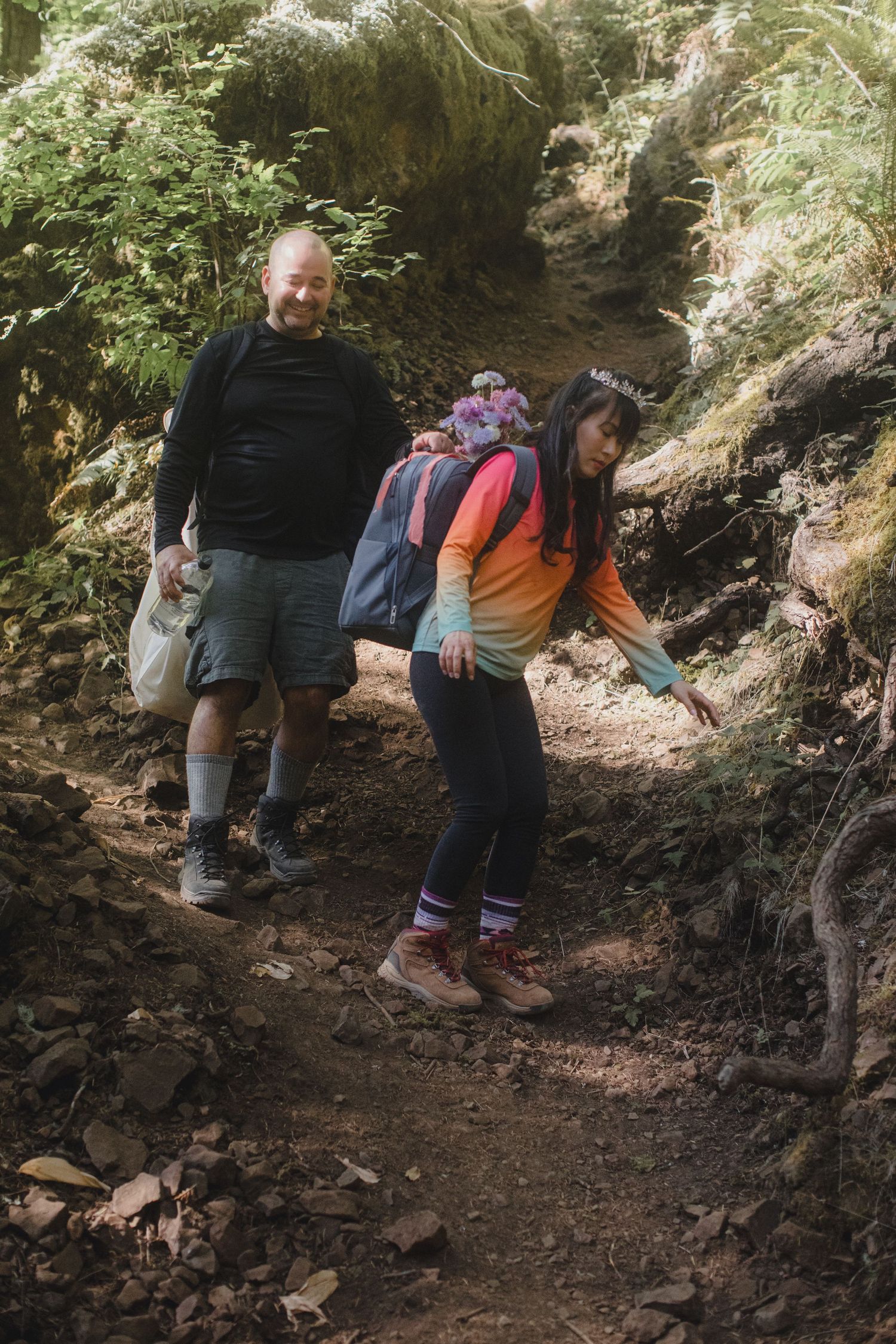 A couple hikes a forest trail, woman in colorful jacket with backpack and man in black shirt carrying a bag.
