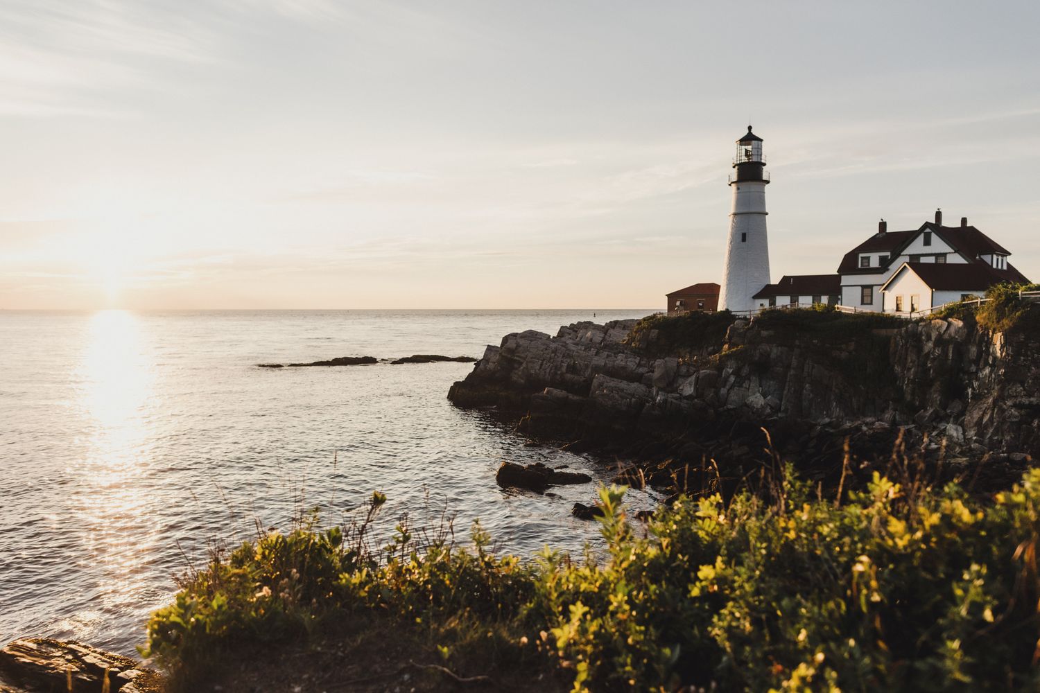 Scenic lighthouse perched on rocky coastal cliffs at sunset with golden wildflowers in the foreground.