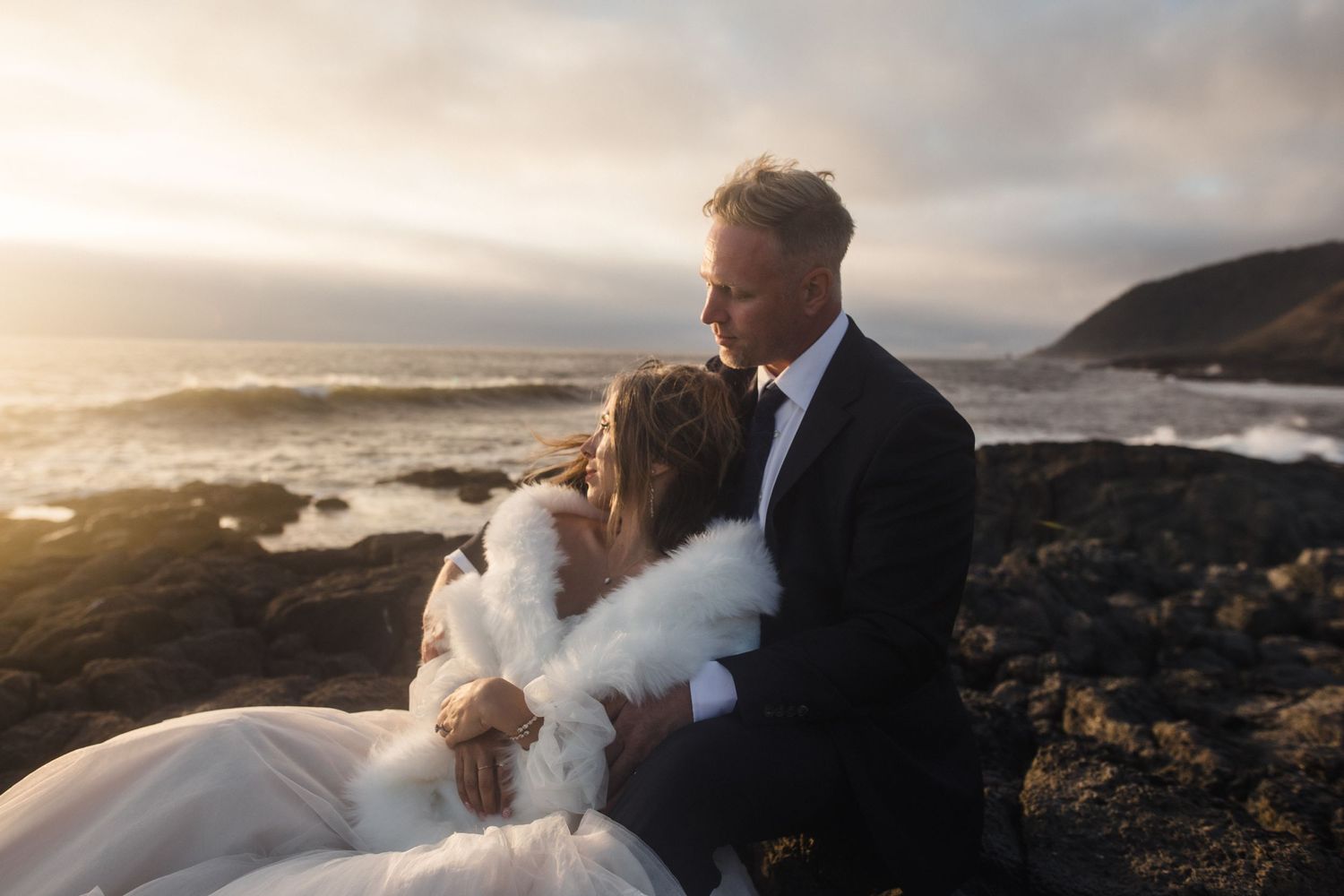 An eloping couple embraces on rocky coastal cliffs on the Oregon Coast during sunset with ocean waves crashing behind them.