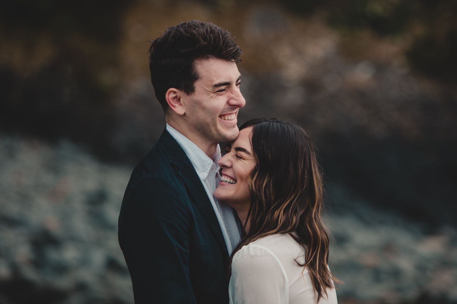 Happy couple embracing outdoors, smiling woman leaning into man in dark blazer with rocky coastal backdrop.