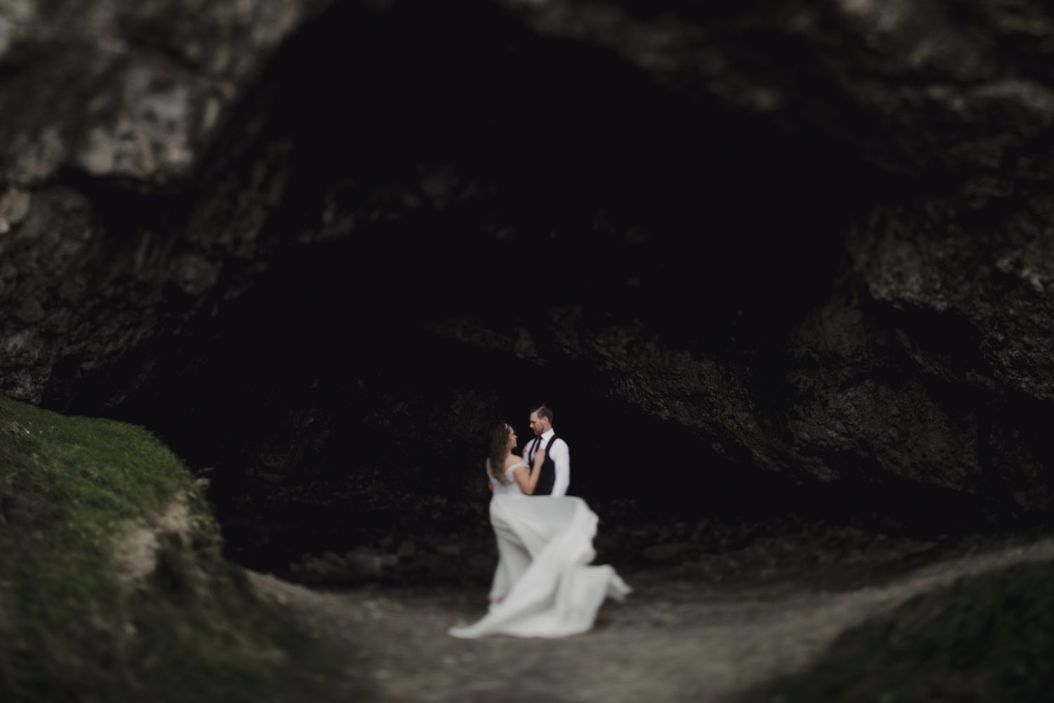 A wedding couple poses together in a dramatic cave entrance with moody lighting.