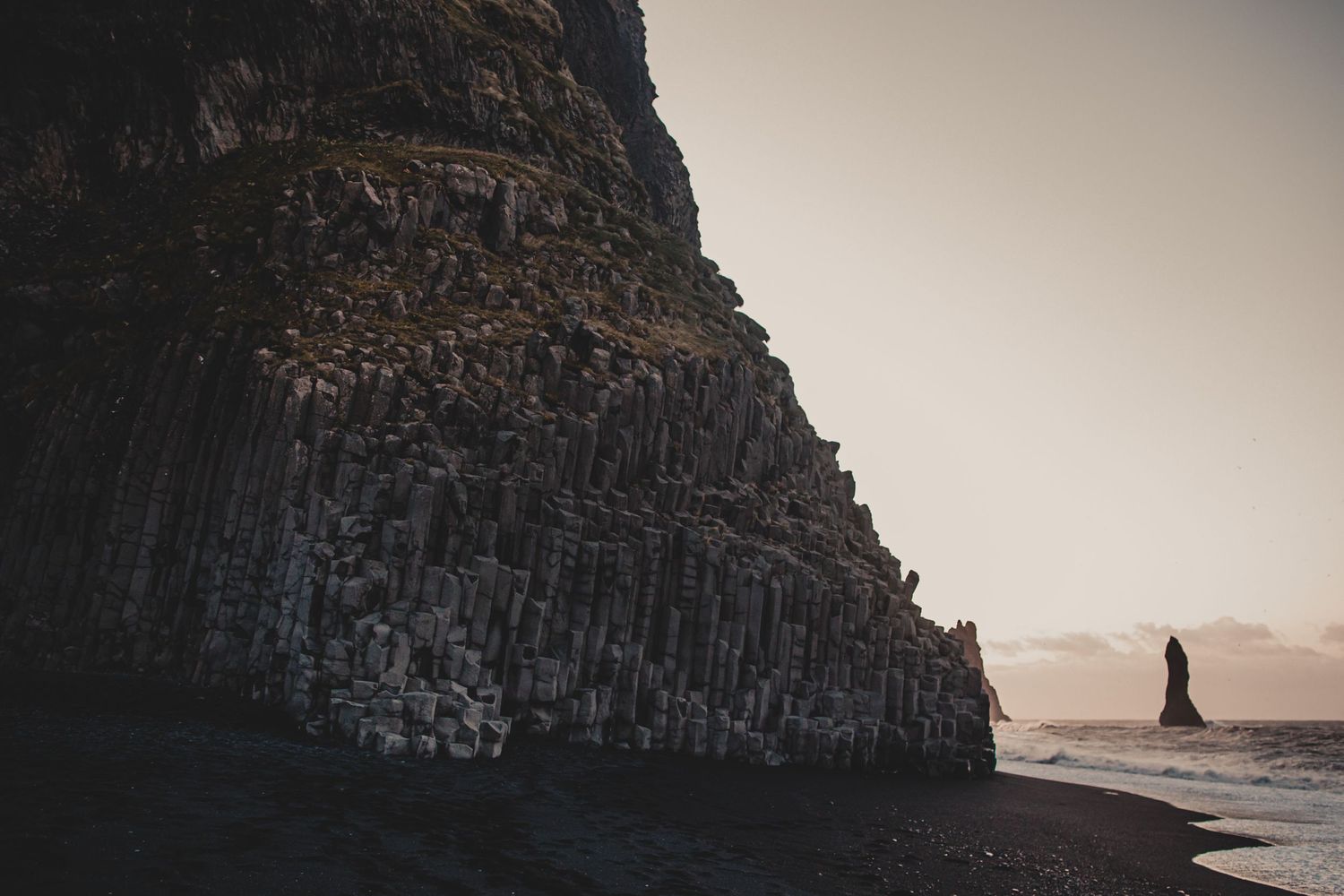 A dramatic basalt cliff formation towers over a black sand beach at sunset.