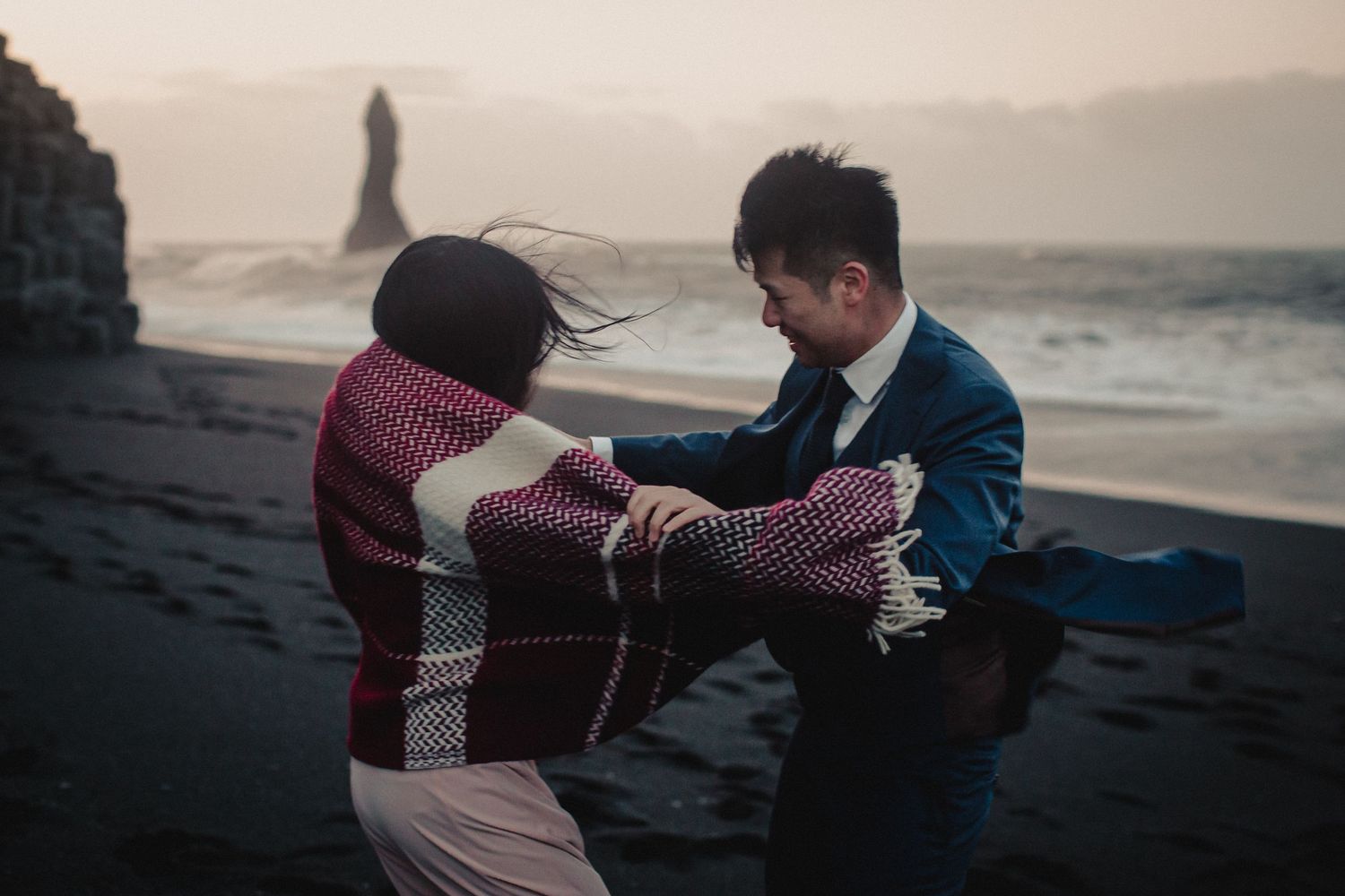 A romantic couple dancing on a black sand beach at sunset with dramatic cliffs in the background.