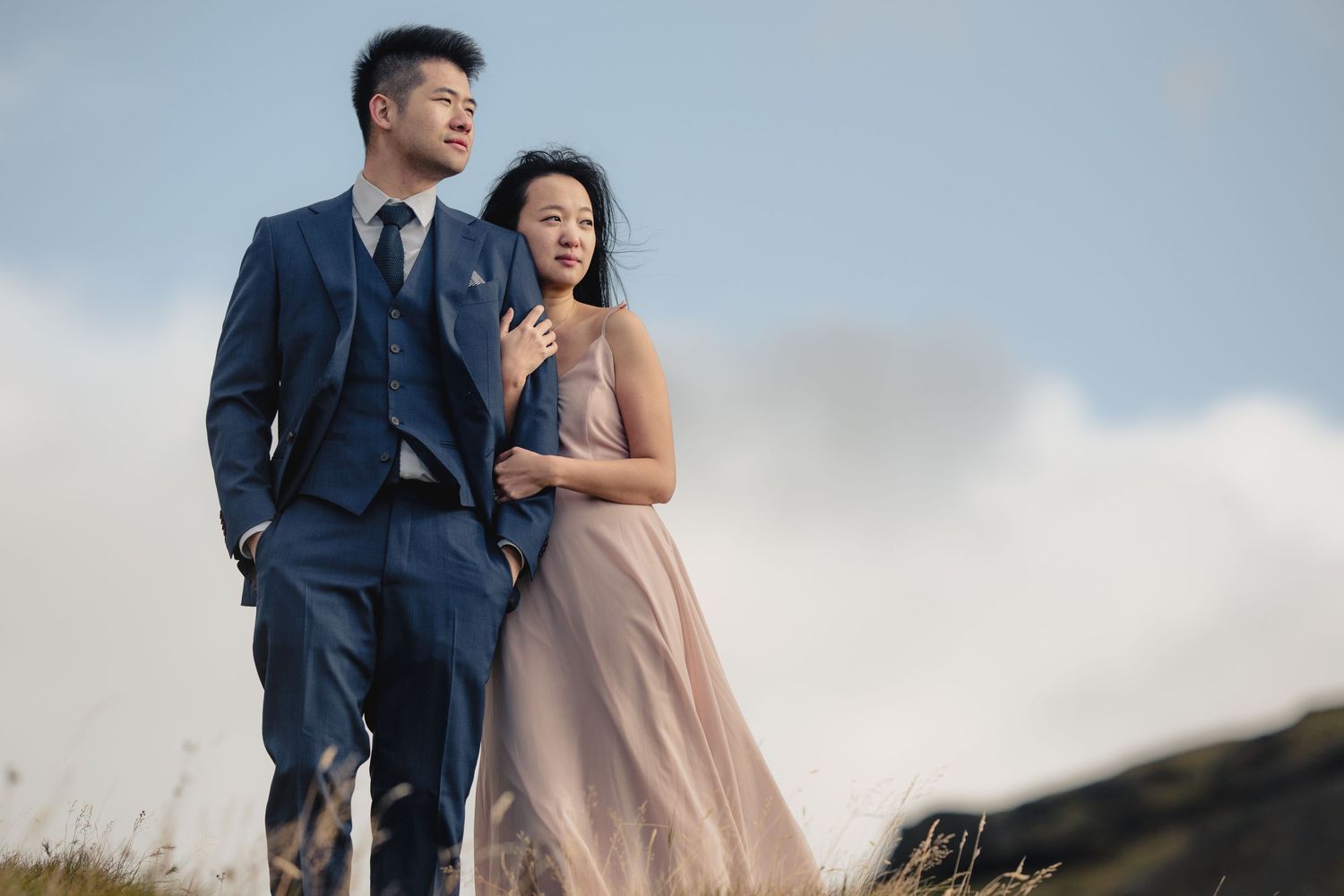 Couple in formal attire embracing on a hilltop against cloudy skies.